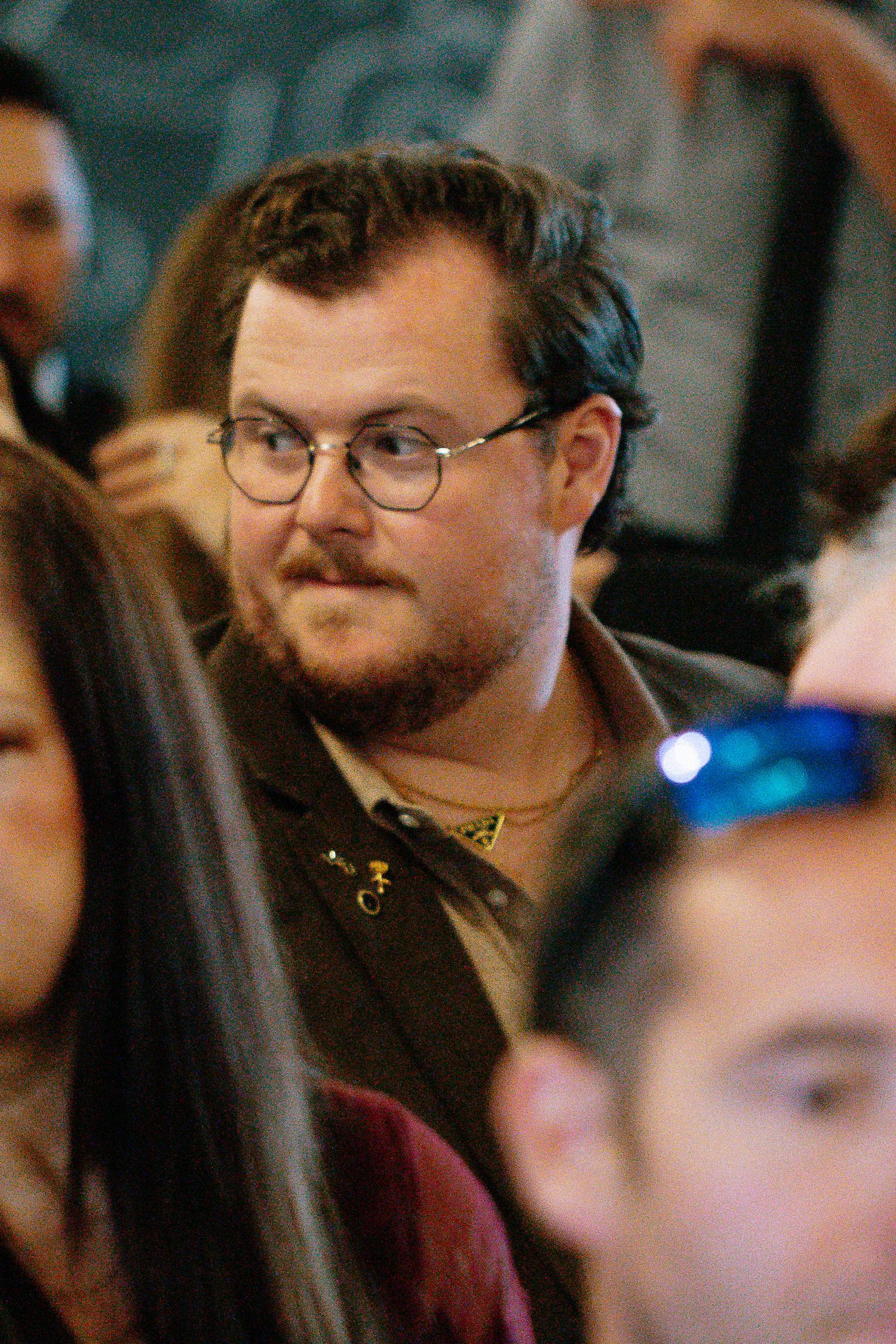 Male wedding guest with glasses and gold chain seated among crowd during wedding ceremony.