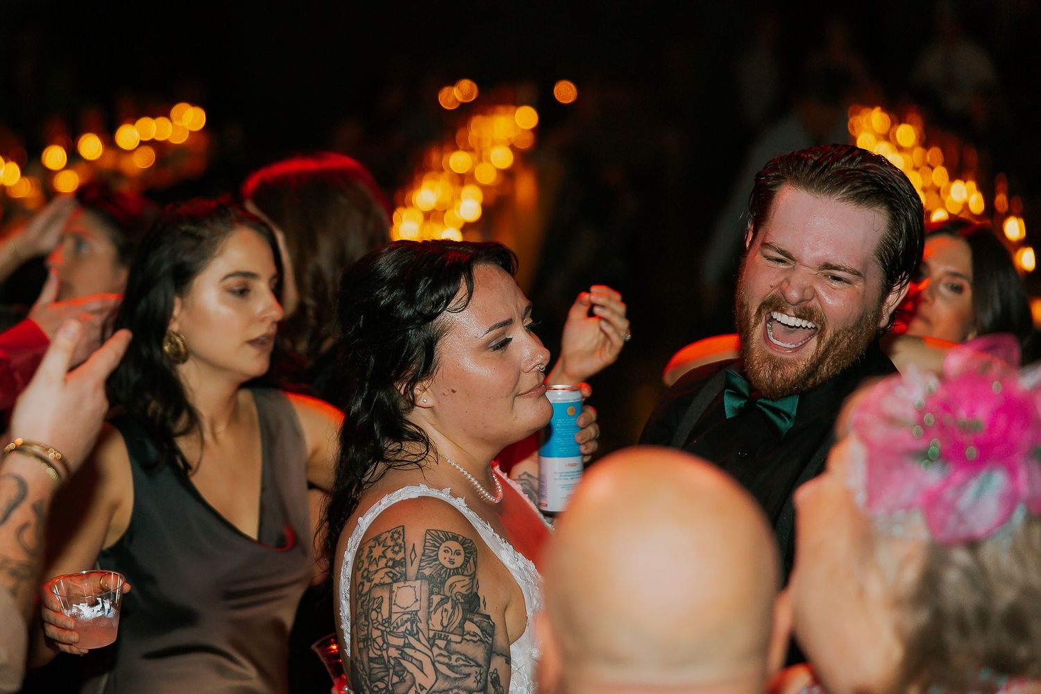 Bride and groom laughing together surrounded by guests on a warmly lit dance floor.