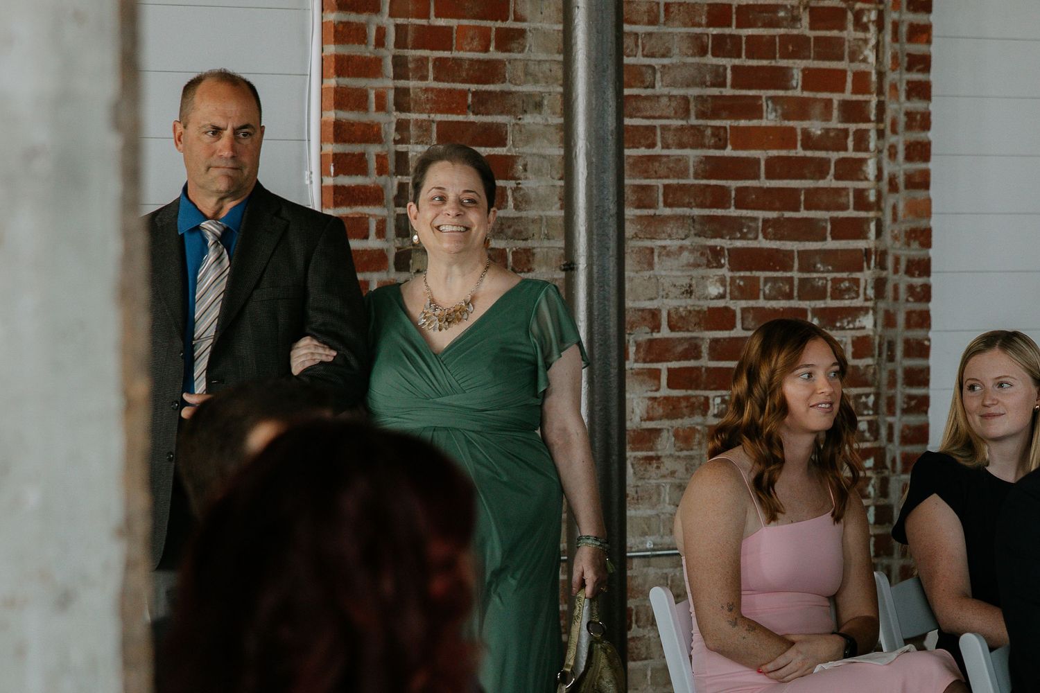 Couple in formal attire stands watching wedding ceremony in rustic brick-walled venue.