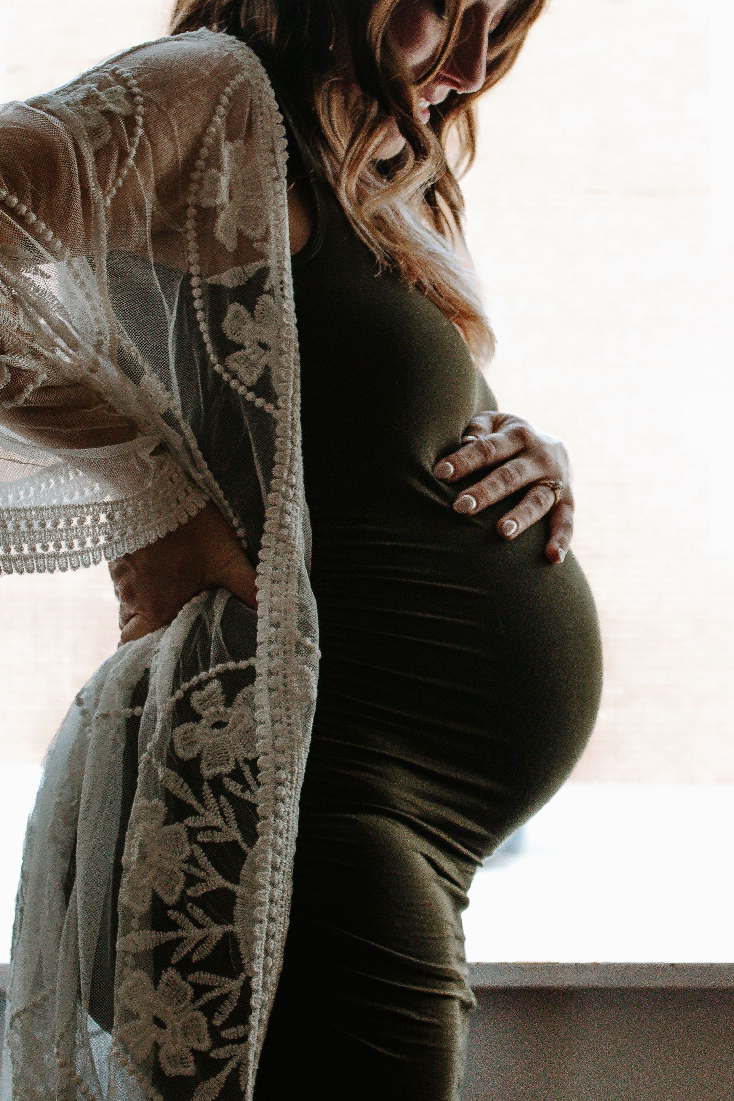 Pregnant woman in olive dress cradling baby bump by window in Cincinnati, Ohio.