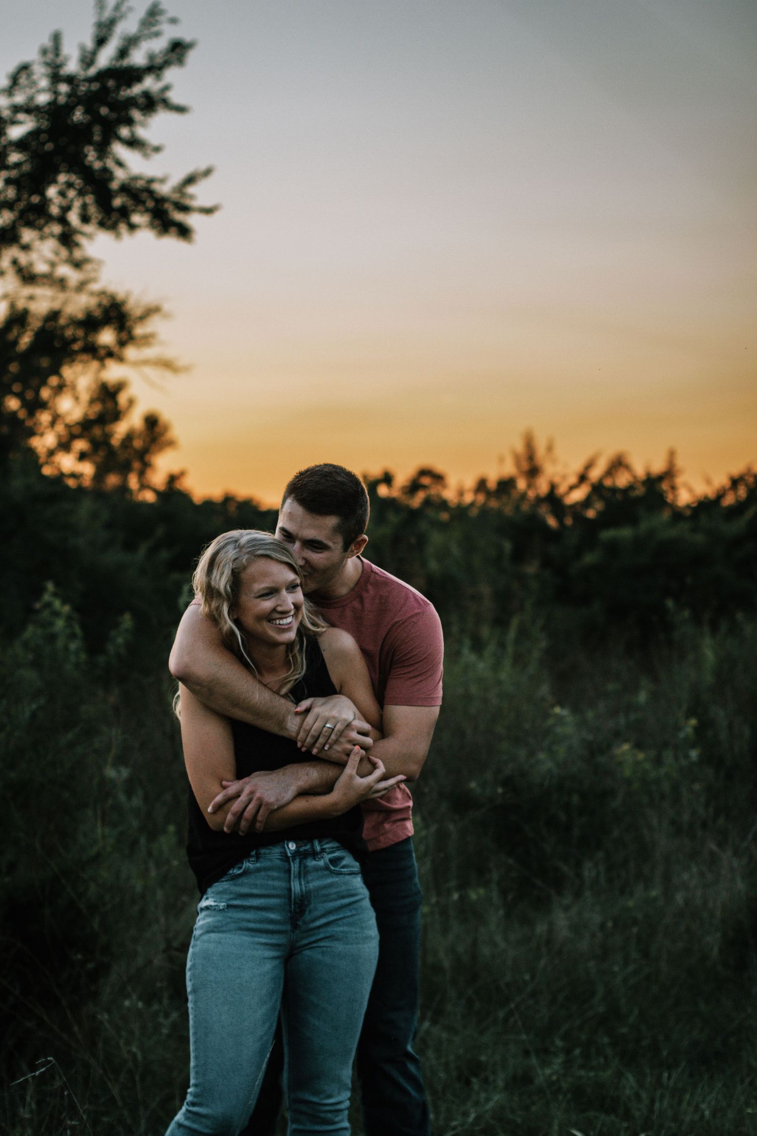 Man hugging woman from behind at sunset in a lush green field, both laughing joyfully in Cincinnati, Ohio