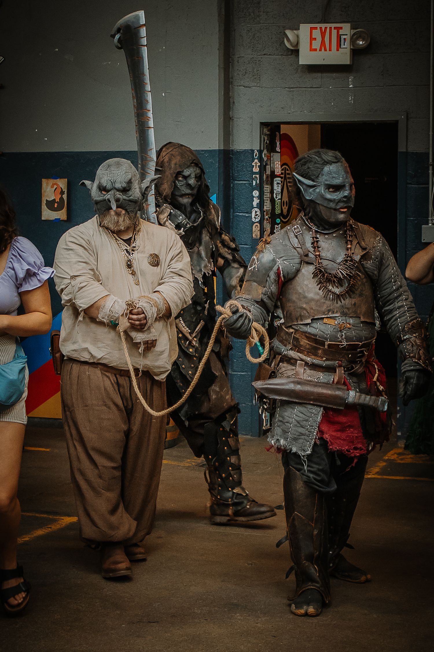 Three people in elaborate orc costumes with detailed prosthetics and armor at a cosplay convention near an exit sign.