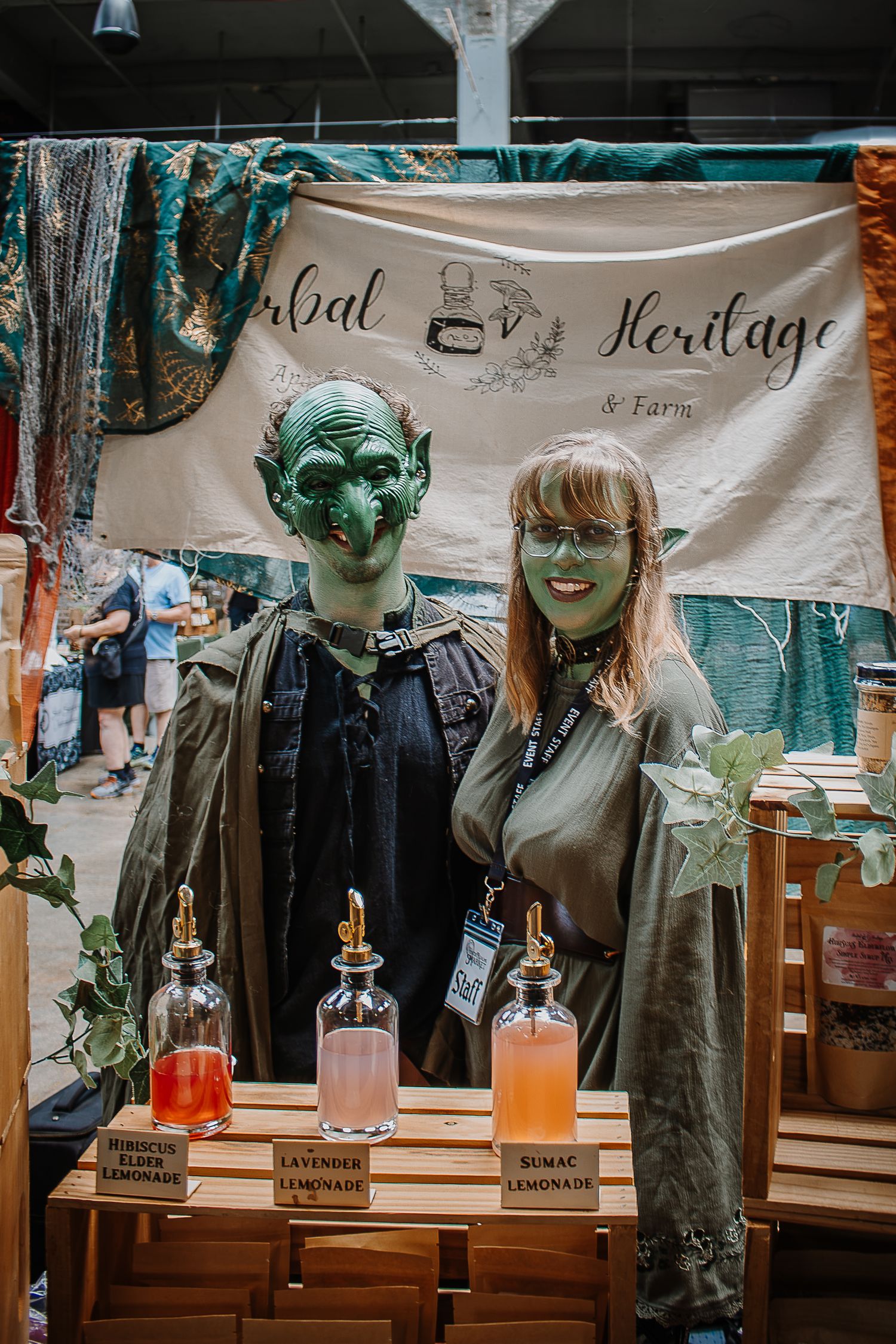 Two people in green goblin makeup pose at Herbal Heritage Farm booth with elderflower, lavender, and sumac lemonades.