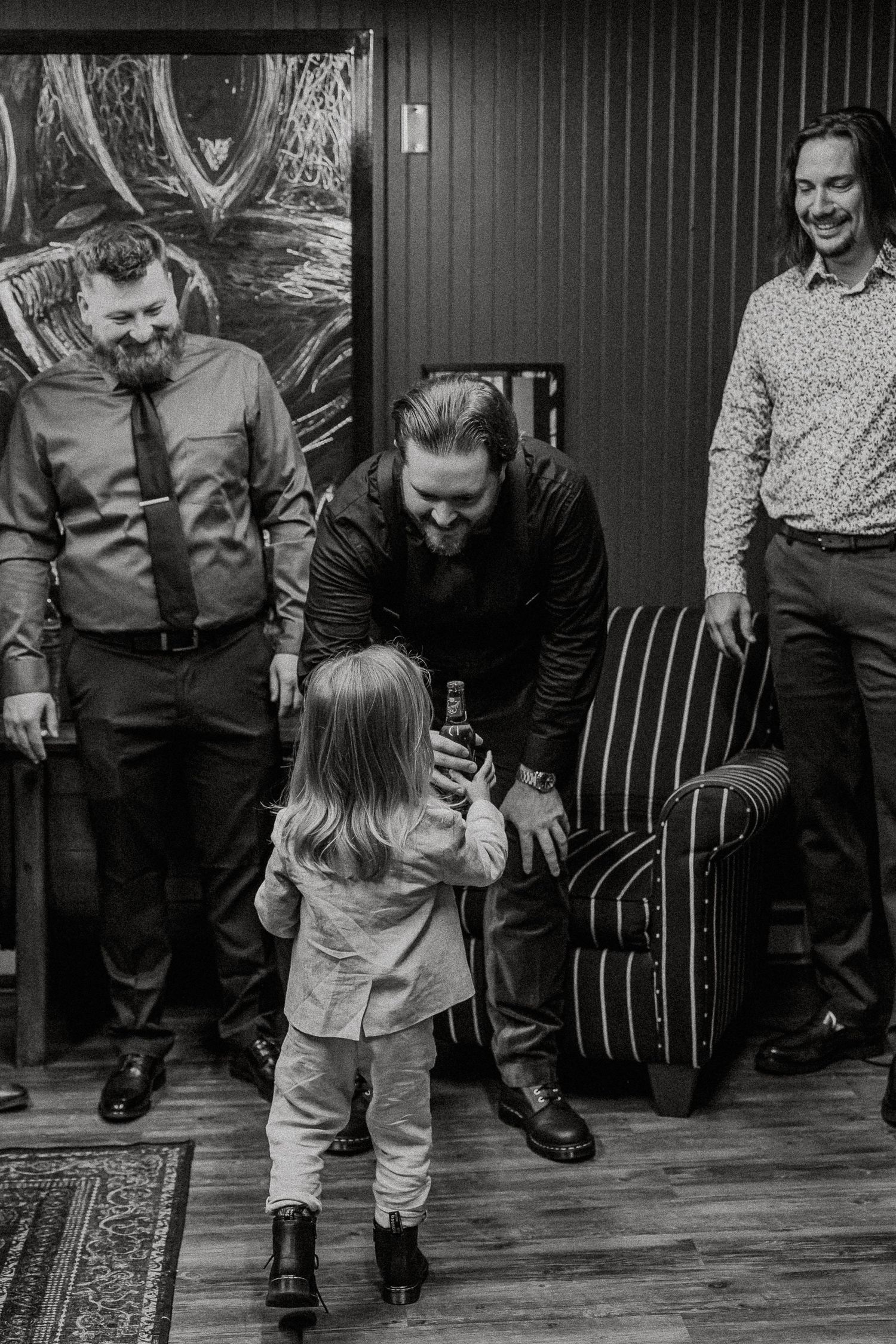 Black and white photo of groom greeting small child with smiling groomsmen in background.