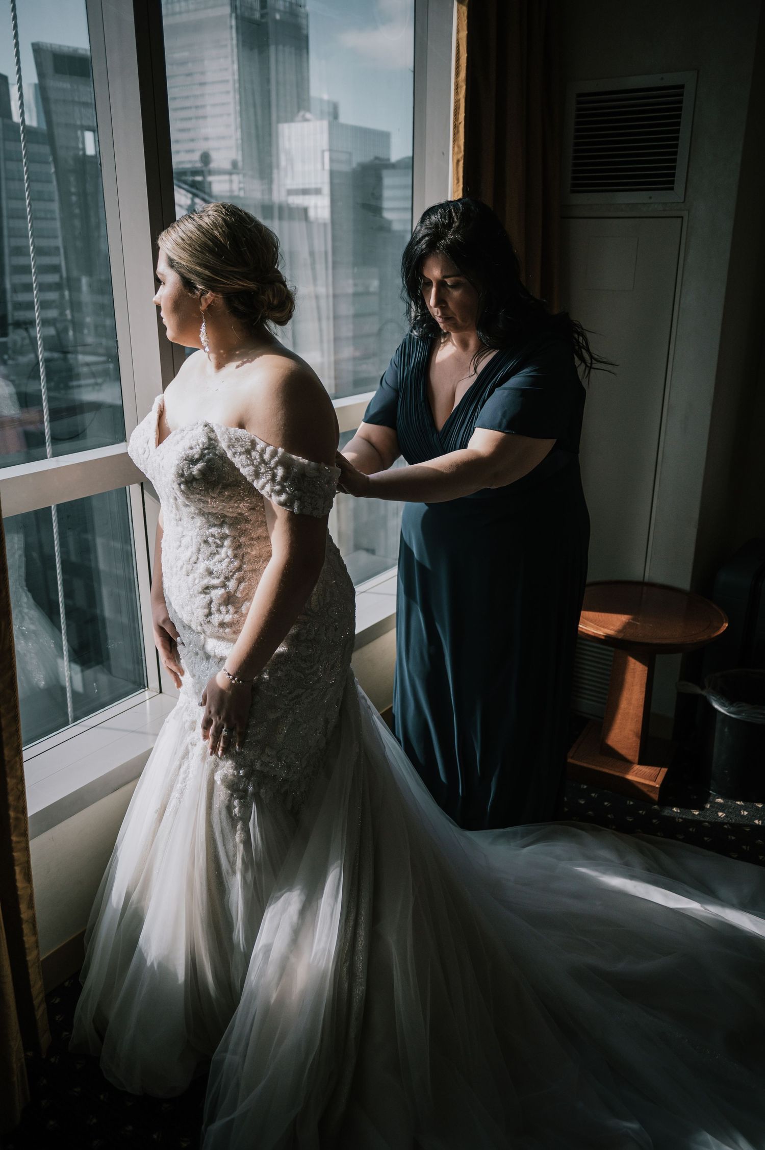Assistant helps bride in lace mermaid gown with buttons by large hotel window overlooking city.