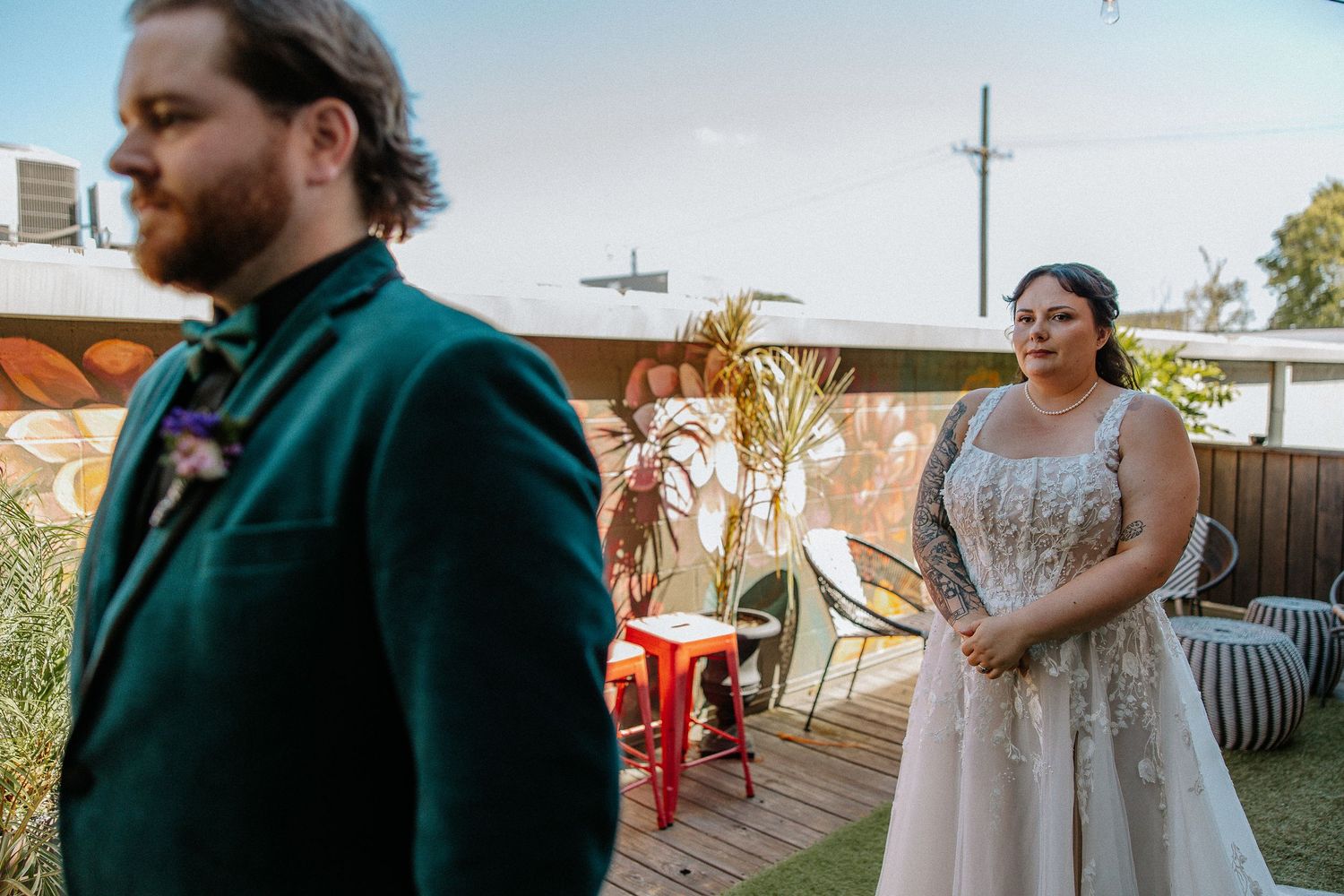 Bride in embellished wedding gown watches groom in emerald velvet suit on rooftop venue during first look.