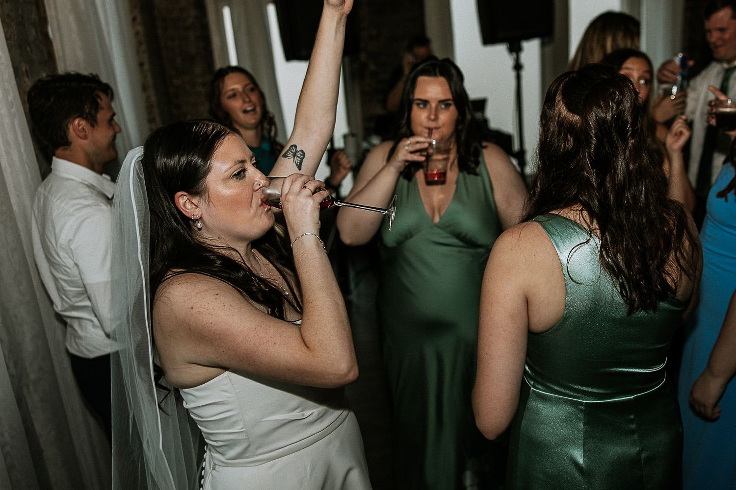 Bride in white gown dances energetically with bridesmaids in sage green dresses at wedding reception.