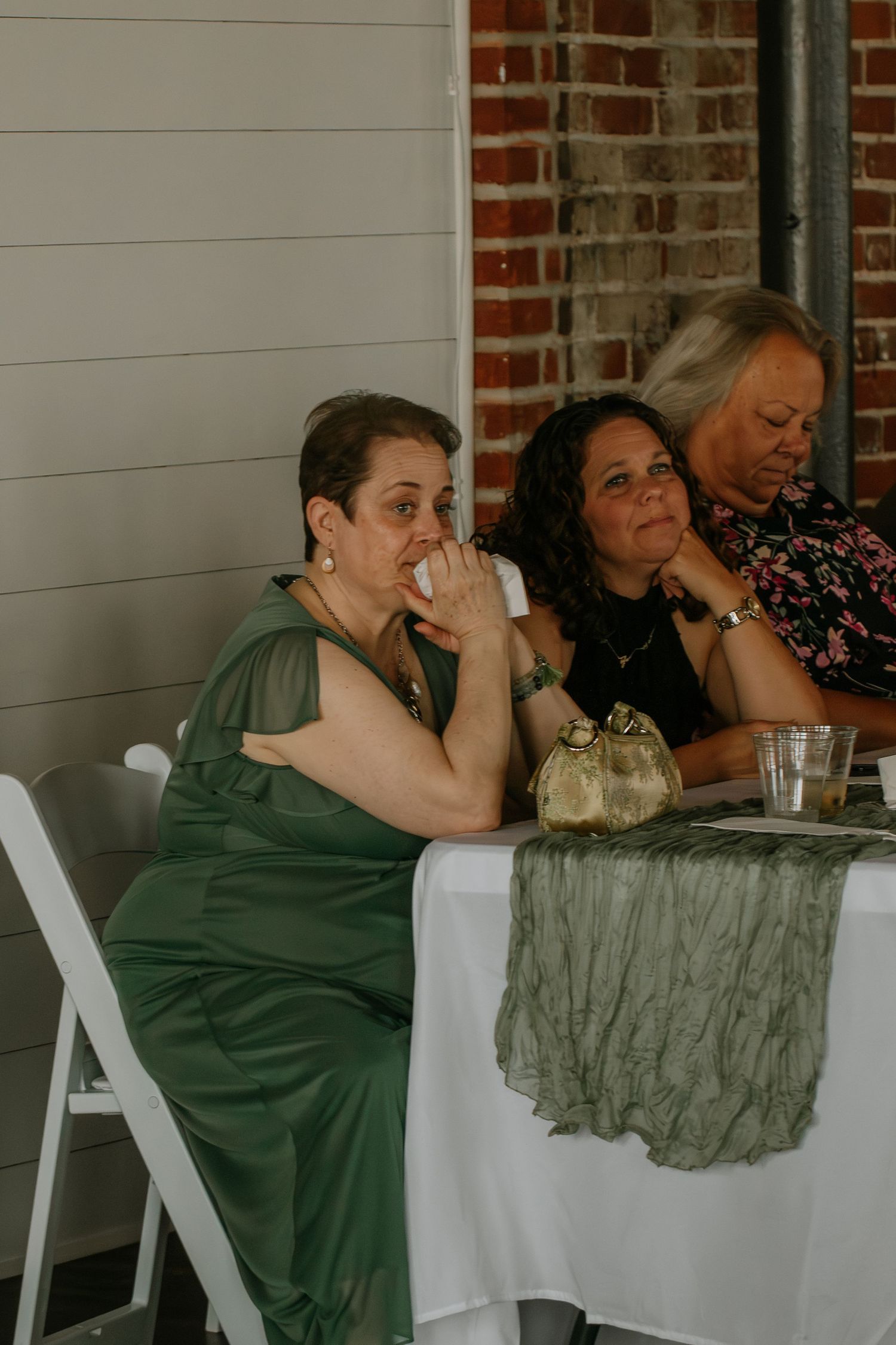 Wedding guests in green dresses sit at a white-draped table during an outdoor reception ceremony.