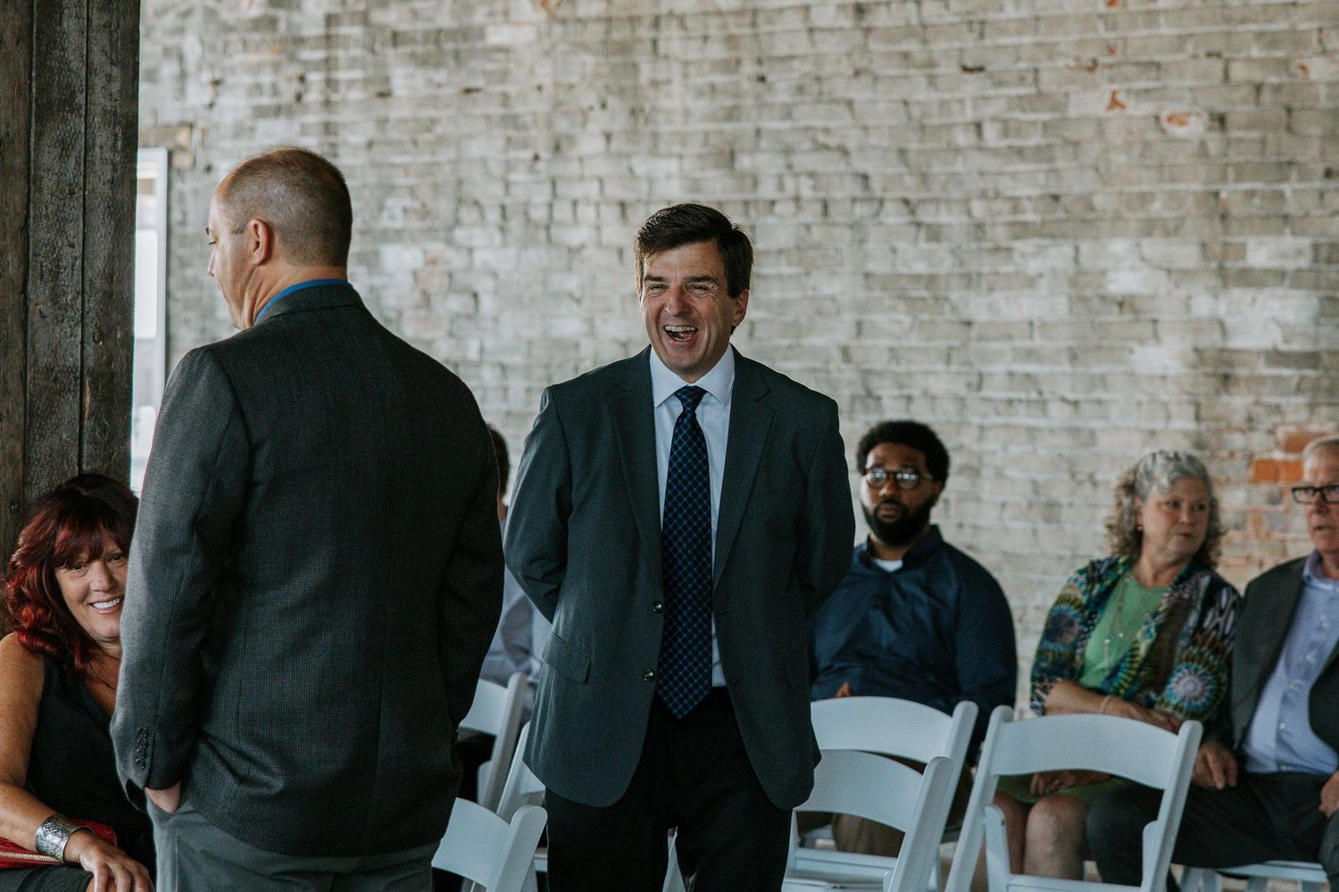 Two men in suits greeting each other at a wedding ceremony with guests seated in background.