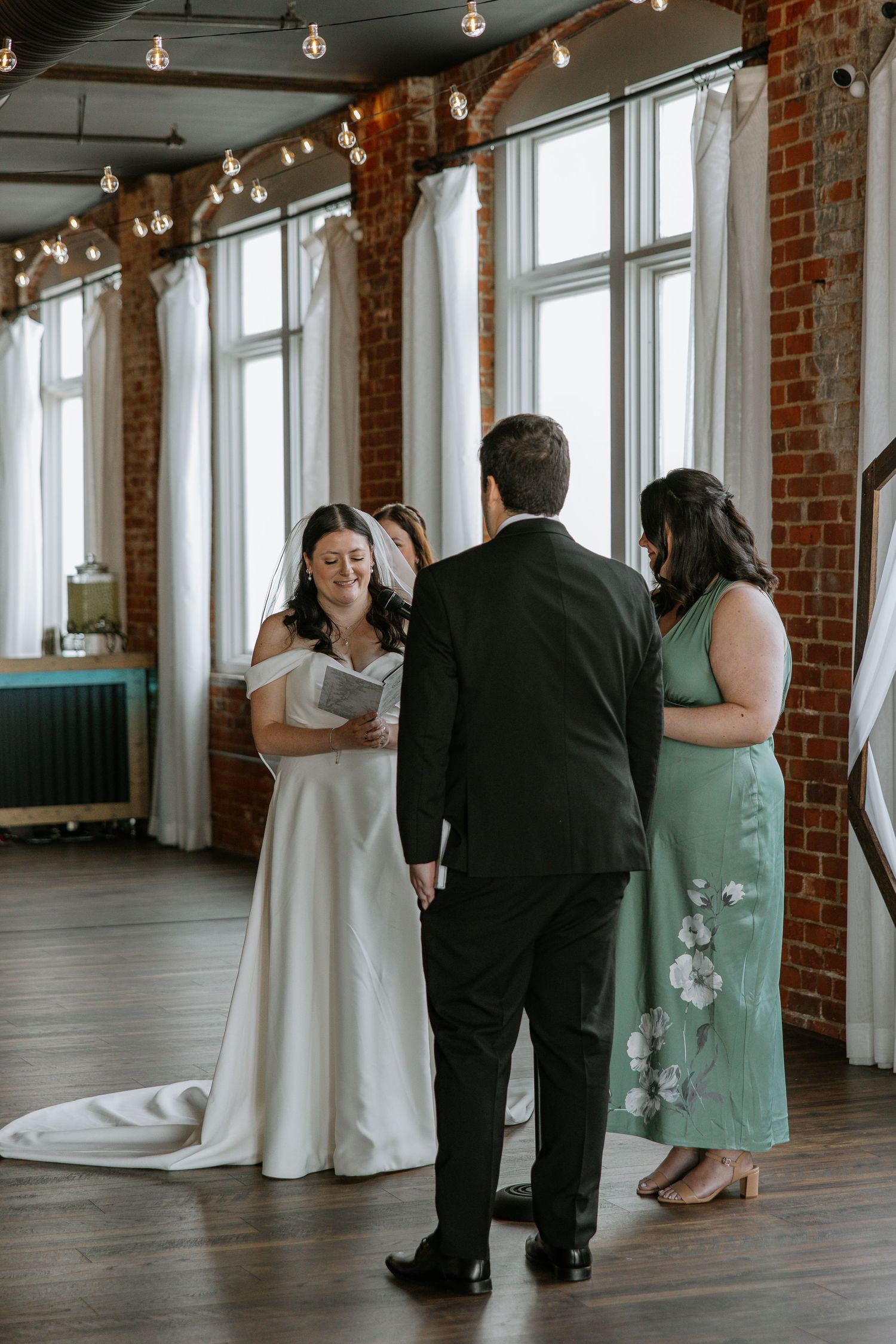 Bride in white gown reads vows to groom in black suit at indoor ceremony.