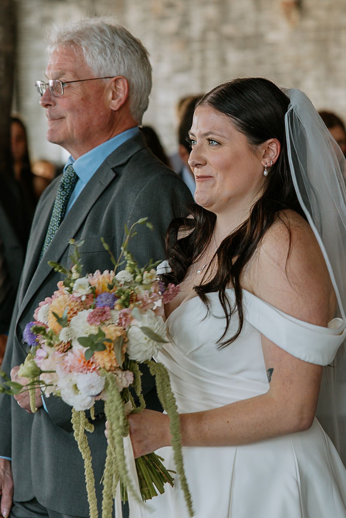 Emotional bride in off-shoulder gown holds colorful bouquet while walking down wedding aisle.