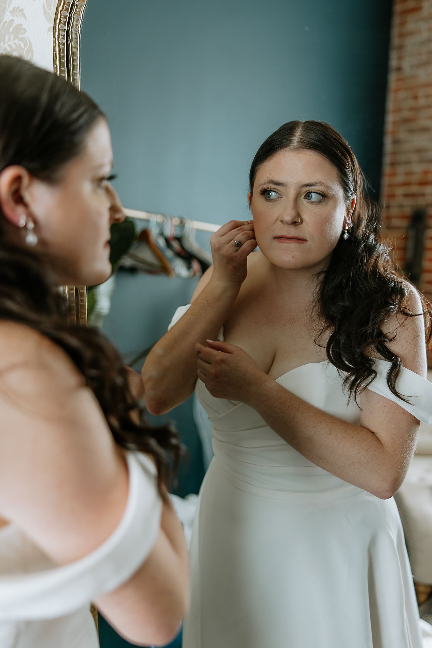Bride putting on earrings while looking at her reflection in an ornate gold mirror.