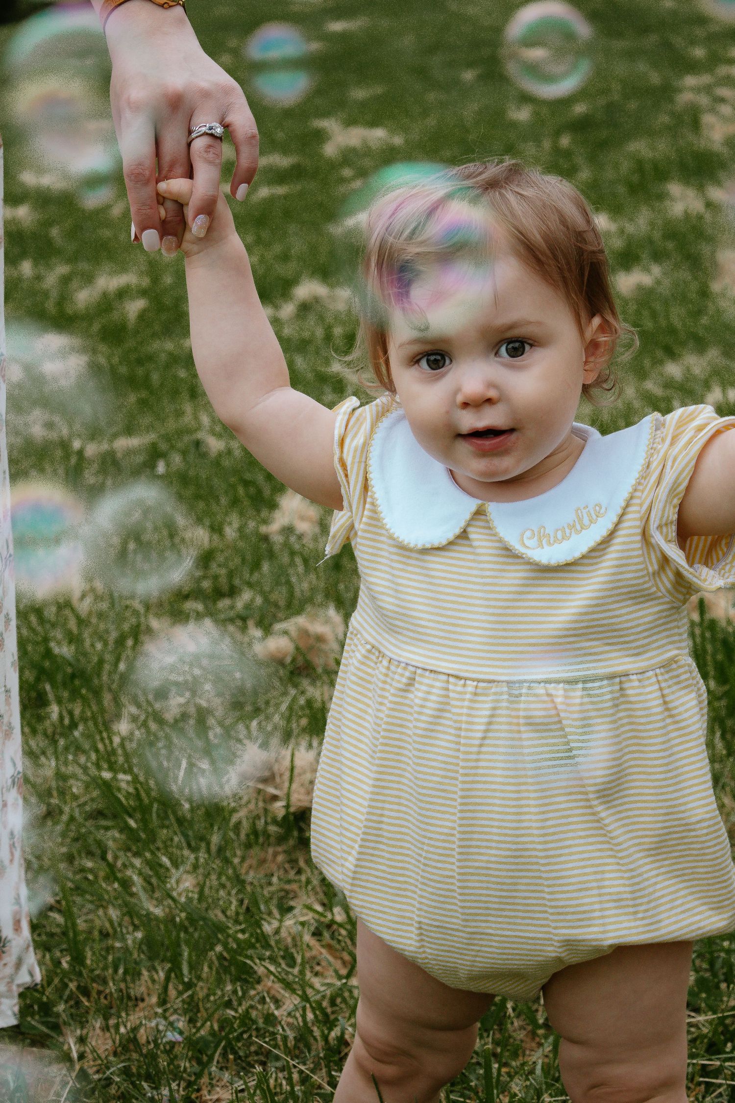 Curious baby girl in monogrammed yellow dress reaching up while surrounded by soap bubbles at French Park in Cincinnati, Ohio