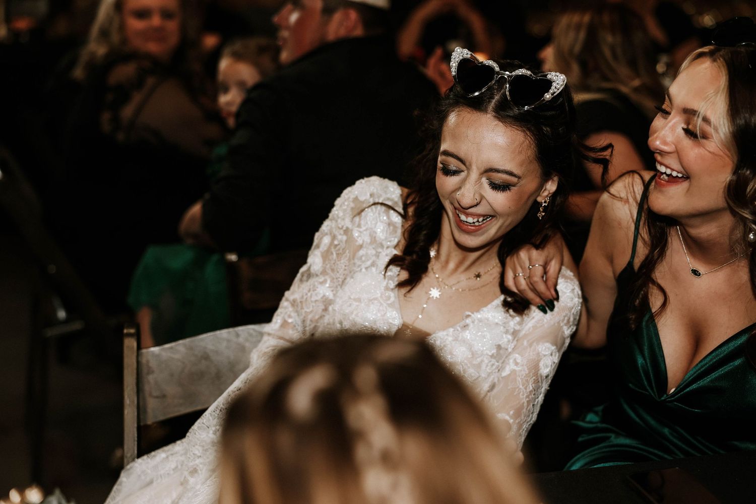 Joyful bride in lace wedding dress laughing with bridesmaid in emerald green dress at wedding reception.