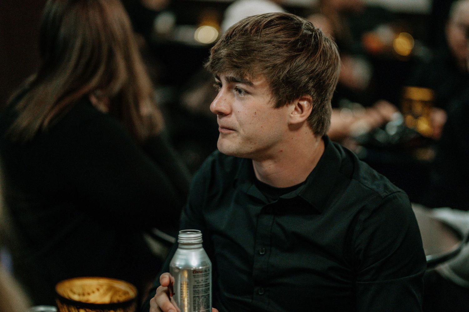 Young man in black shirt sitting at a table holding a water bottle, looking thoughtfully to the side.