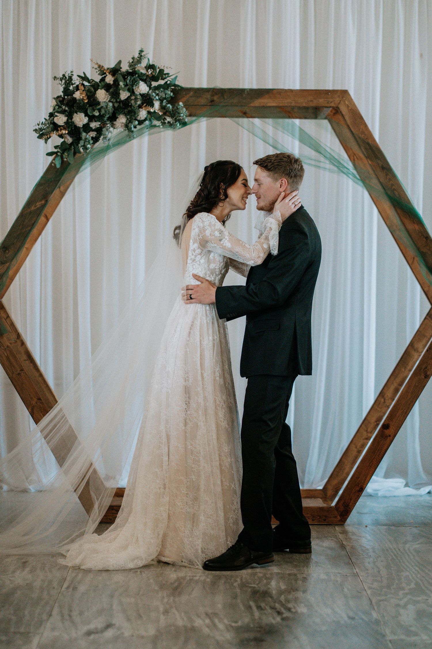 Bride and groom share a romantic moment under a wooden hexagon arch adorned with white flowers and greenery.