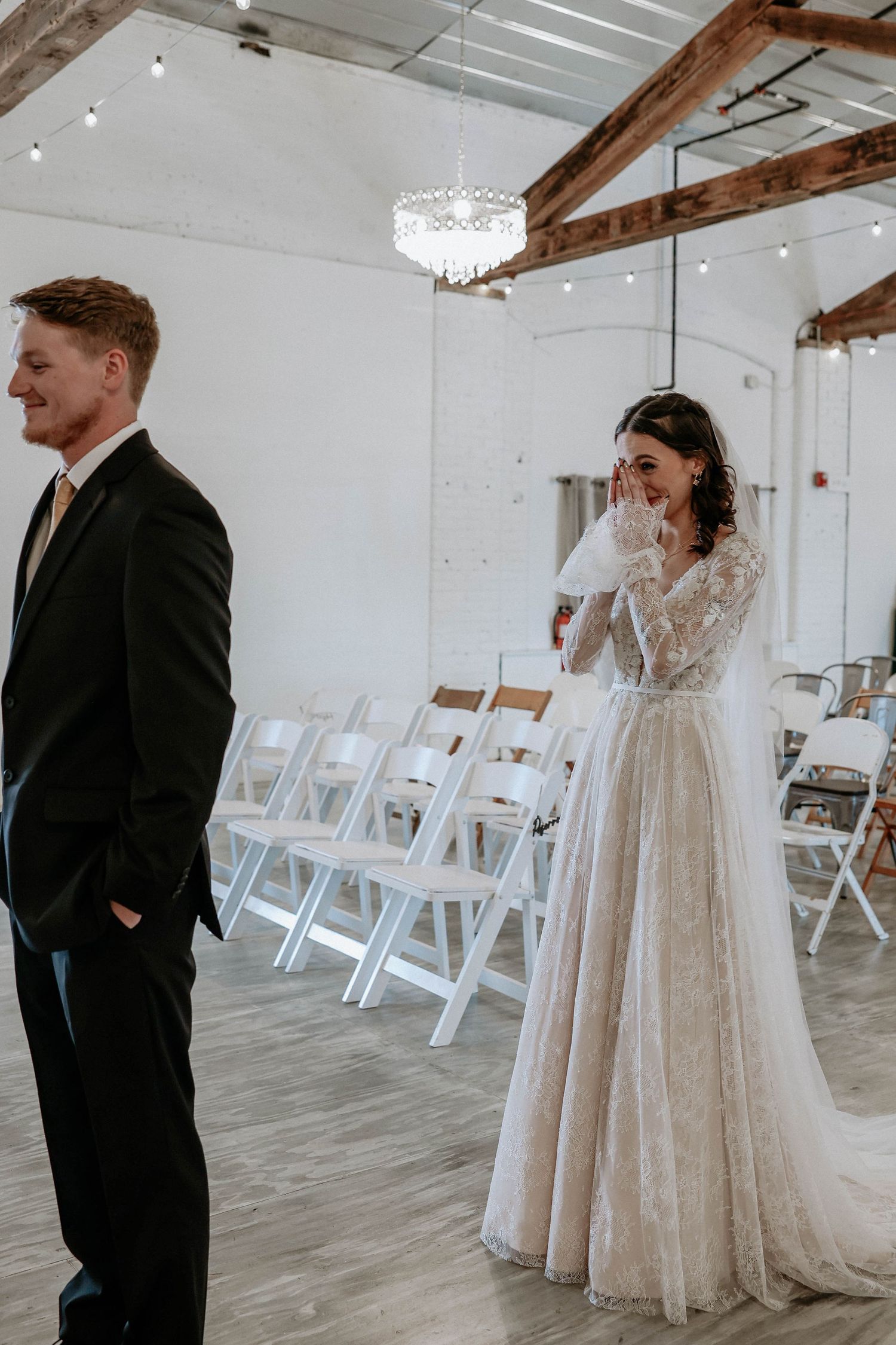 Emotional bride in lace gown covers mouth during first look with groom in black suit at elegant white wedding venue.