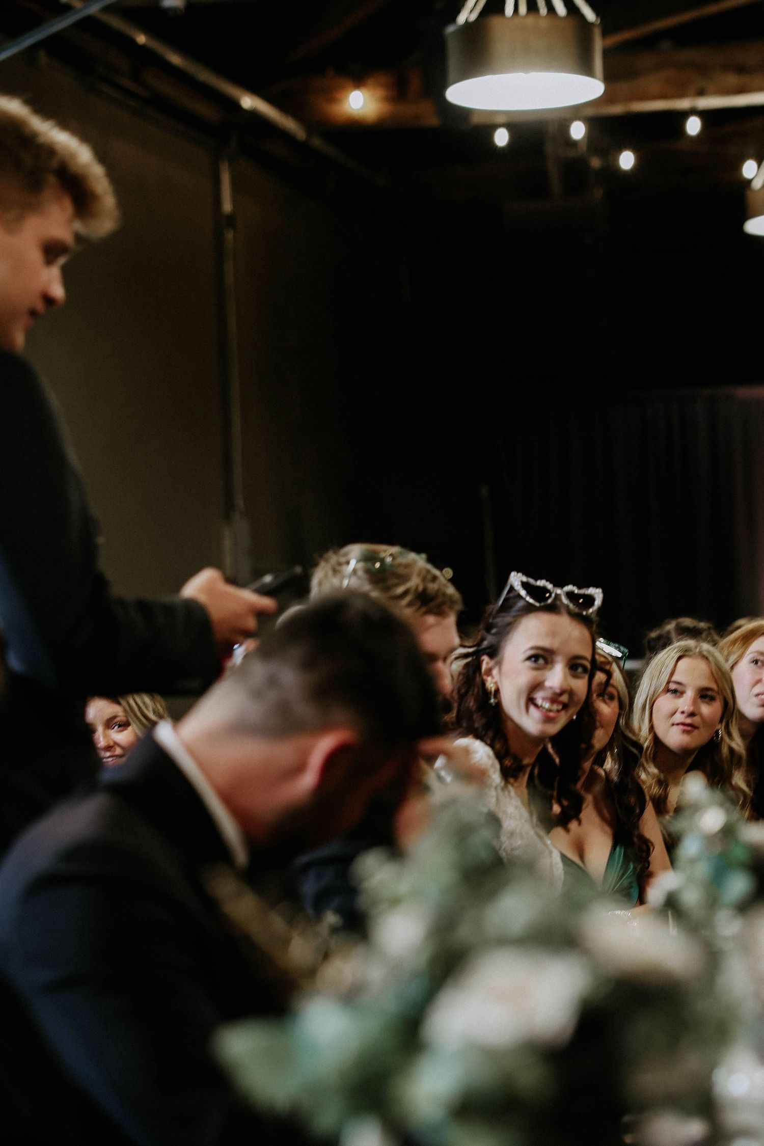 Smiling wedding guests seated at reception tables, woman wearing heart-shaped sunglasses enjoys celebration in dimly lit venue.