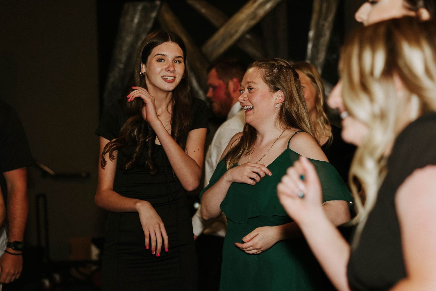 Two women in elegant dresses smile and dance together at a formal evening event or wedding reception.
