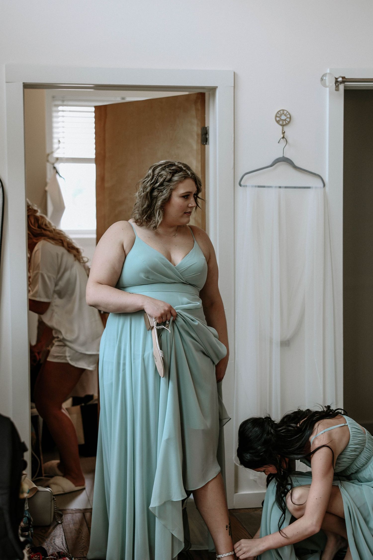 Bridesmaids in sage green dresses getting ready, with white wedding gown hanging in background doorway.