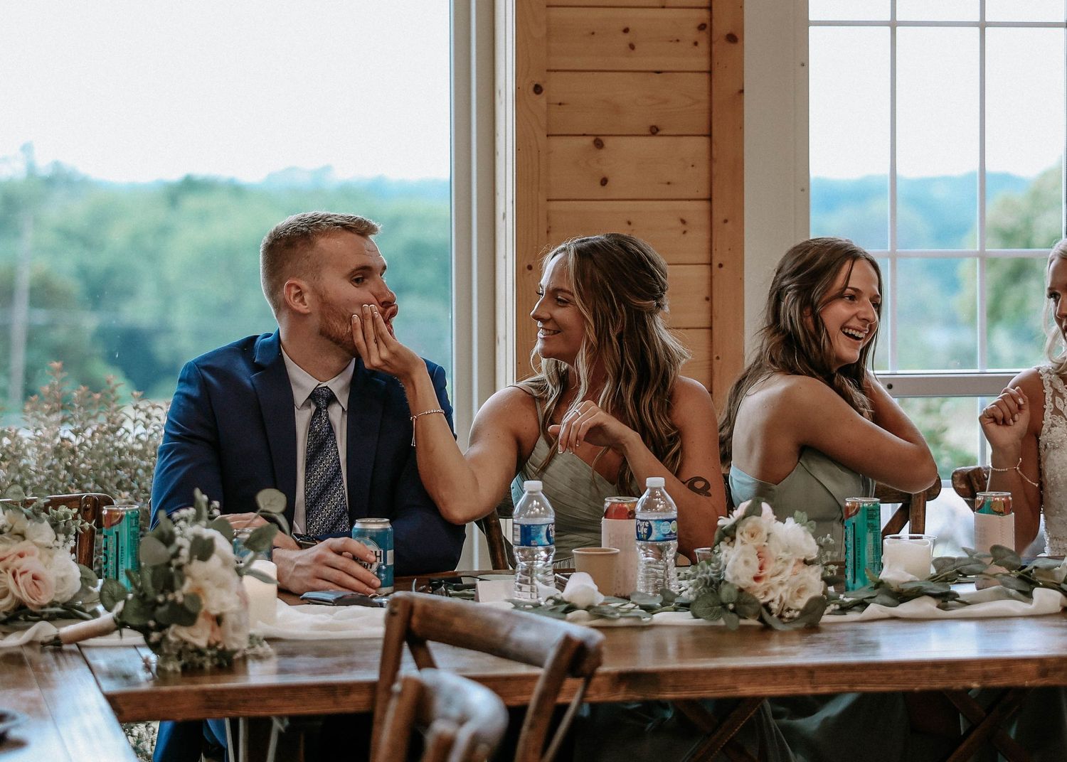 Wedding party laughing joyfully at rustic reception table with floral centerpieces and large windows in background.