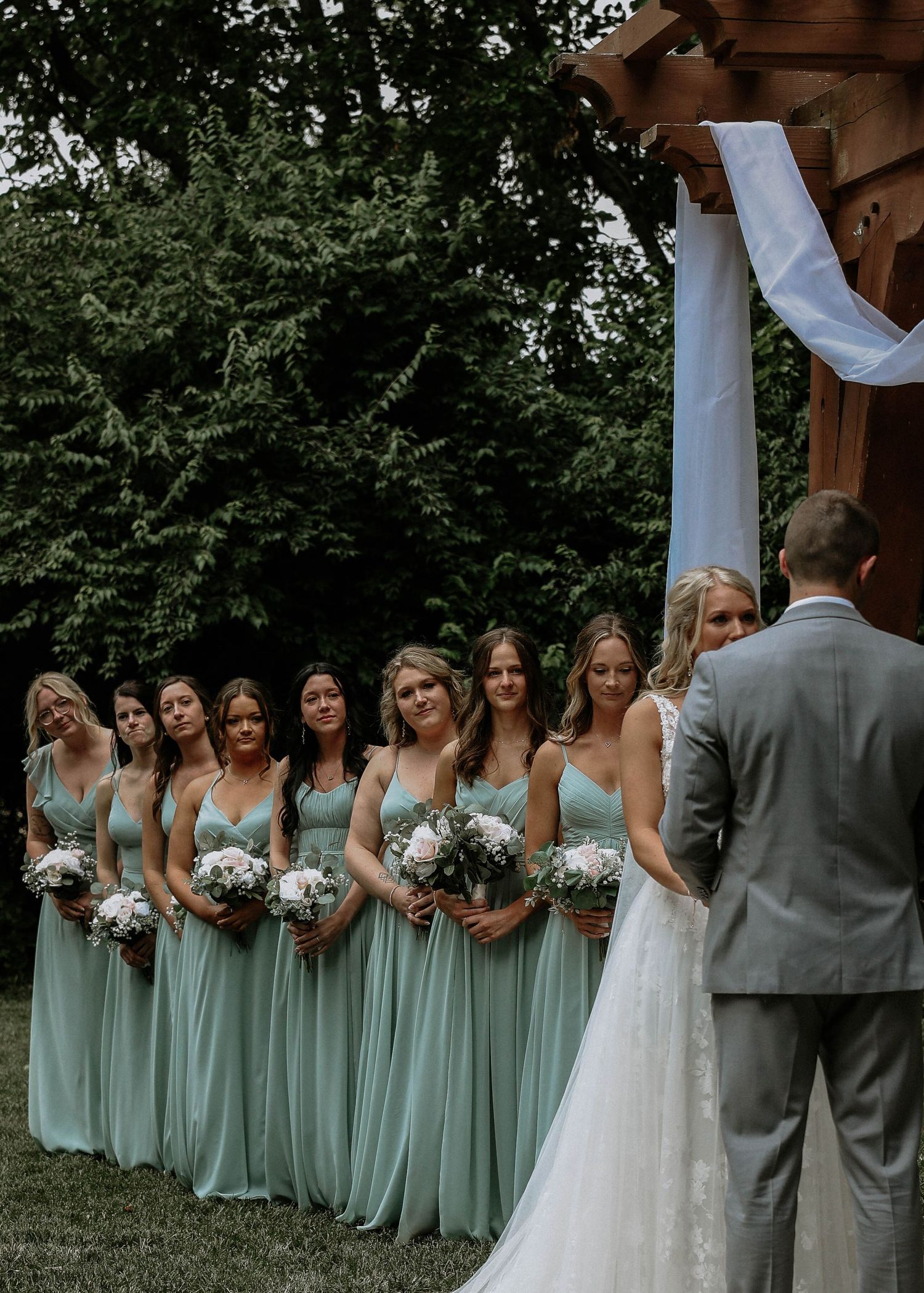 Bridesmaids in sage green dresses holding white bouquets stand beside bride in white gown at outdoor wedding ceremony.
