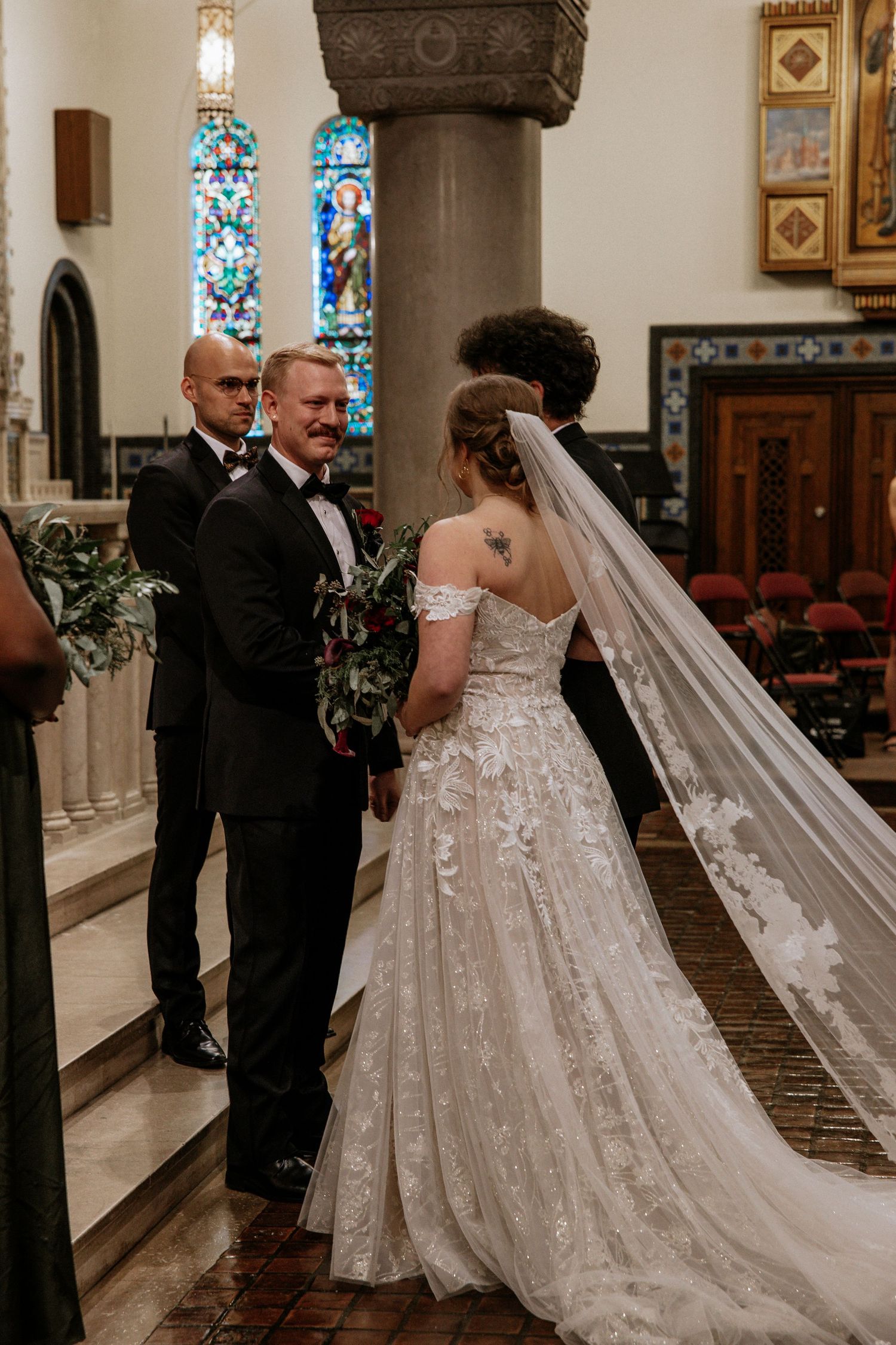 Bride in off-shoulder lace gown with cathedral veil stands at church altar with groom and officiant in Cincinnati, Ohio