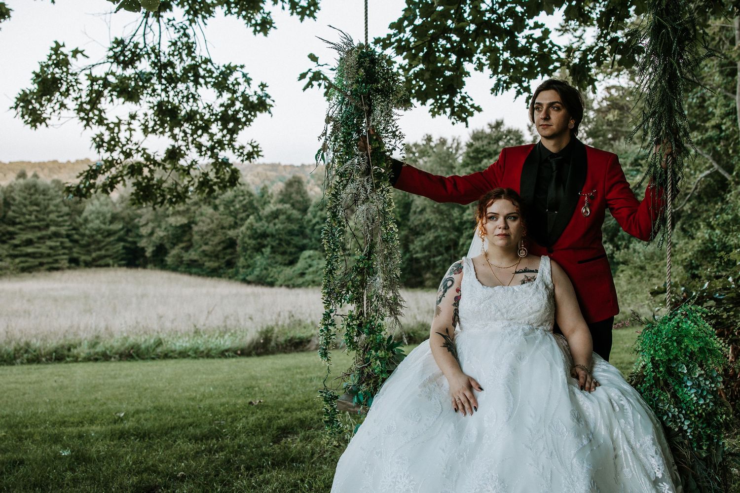 Bride in white ball gown sits on floral swing while groom in red suit stands behind her in lush green outdoor setting.