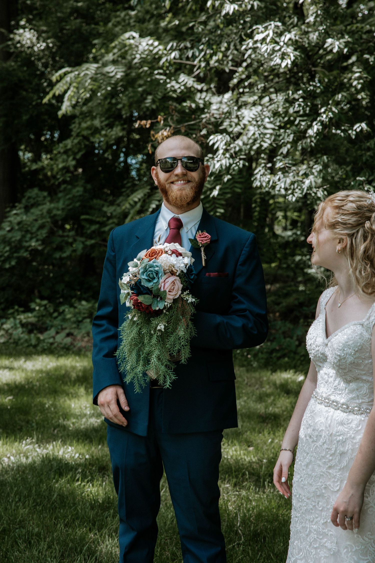 Bearded groom in navy suit wearing sunglasses holds bridal bouquet outdoors in garden at The Barn at Wolf Creek in Akron, Ohio