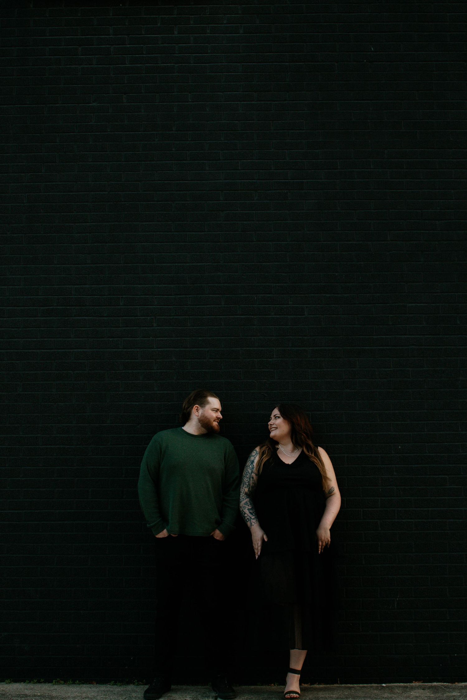 Couple standing apart facing each other against a dark green brick wall in moody lighting in Dayton, Ohio