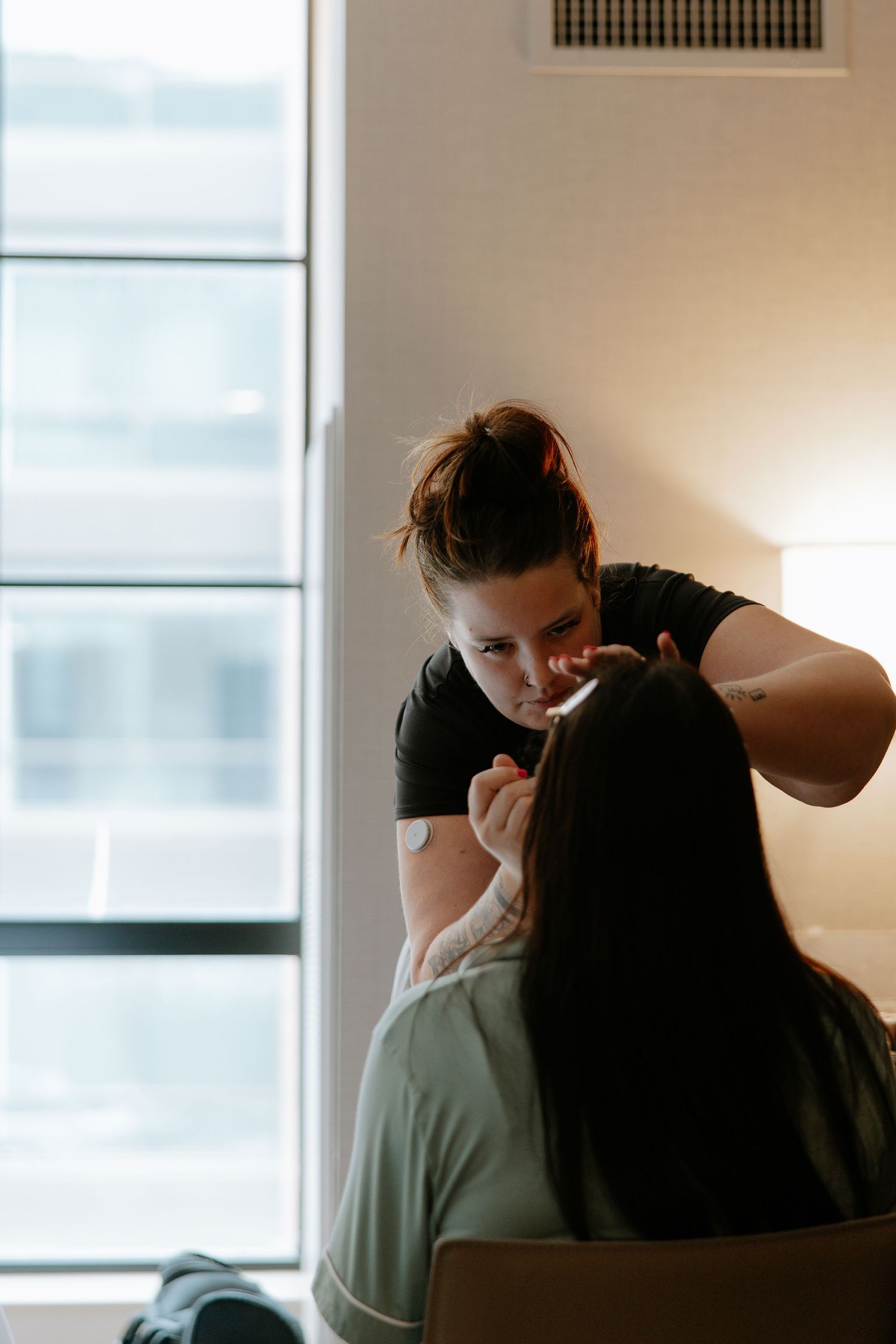 Makeup artist applying bridal makeup to seated woman near bright hotel window.