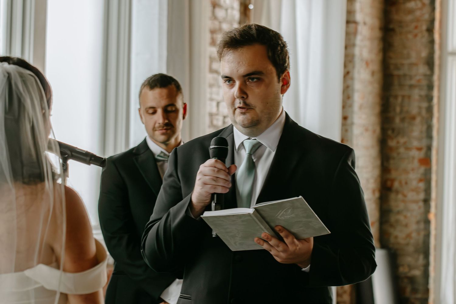 Man in dark suit reading heartfelt speech at wedding ceremony with microphone.