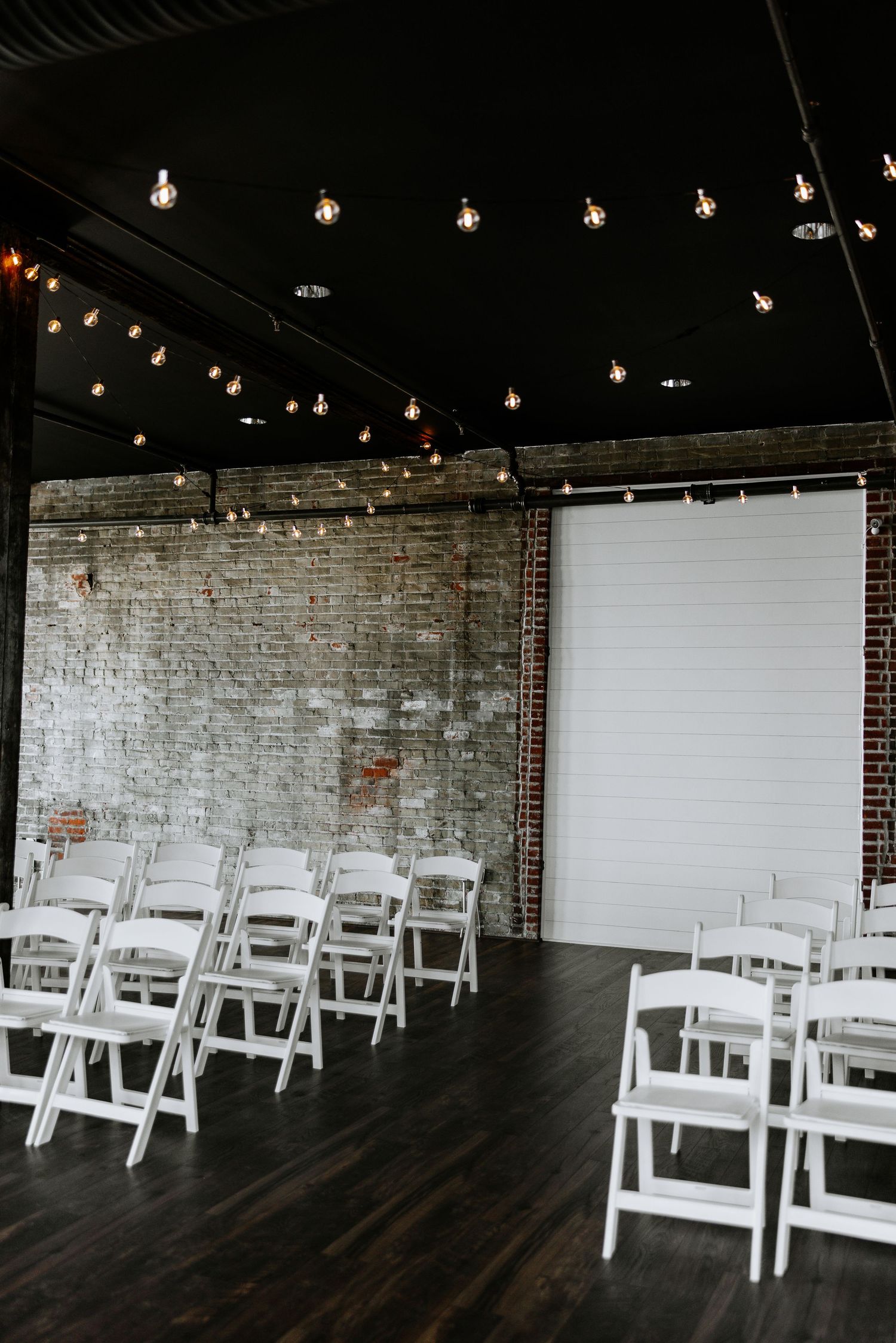 Intimate indoor wedding ceremony venue with white folding chairs, string lights, and exposed brick walls.