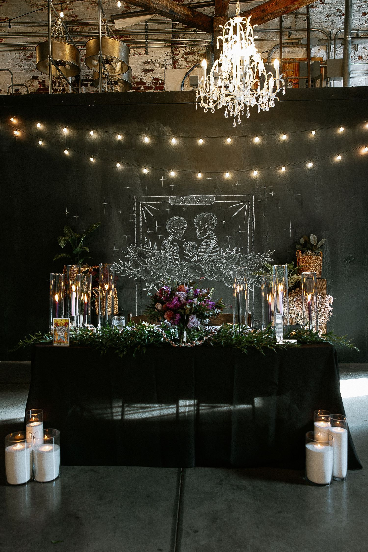 Industrial wedding venue sweetheart table with chalkboard tarot art backdrop, chandelier, and string lights.