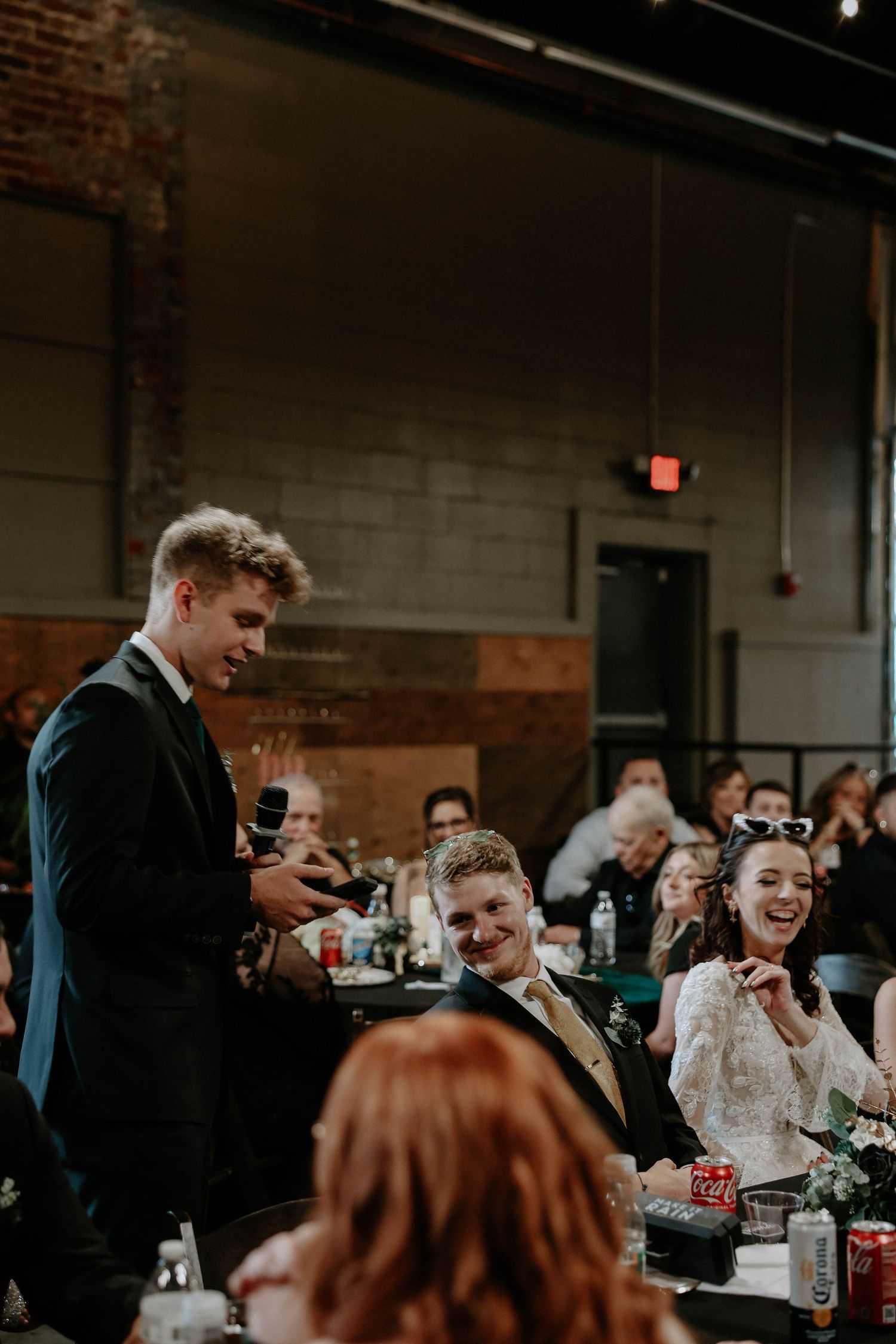 A young man in a suit gives a wedding toast with a microphone while guests laugh and smile at a reception.