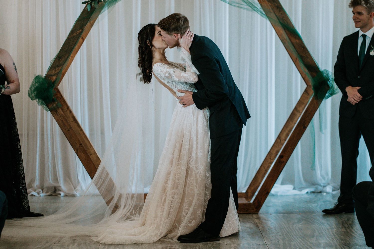 Bride and groom share first kiss under wooden hexagon arch with white draping at wedding ceremony.