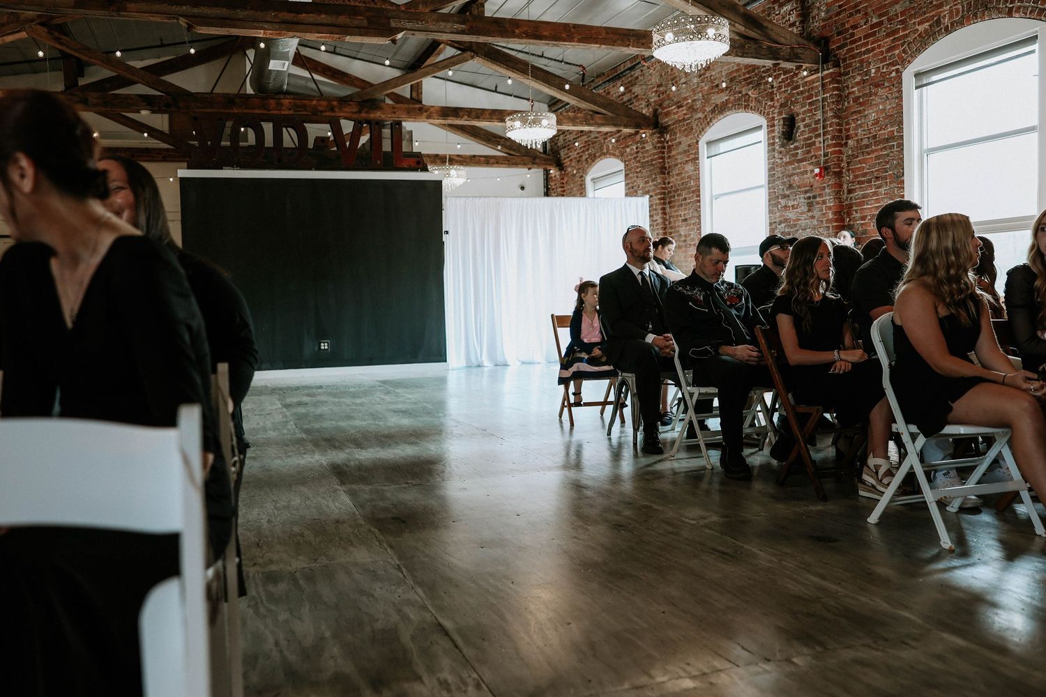 Guests seated in white chairs at an indoor wedding ceremony in a rustic brick venue with hardwood floors.