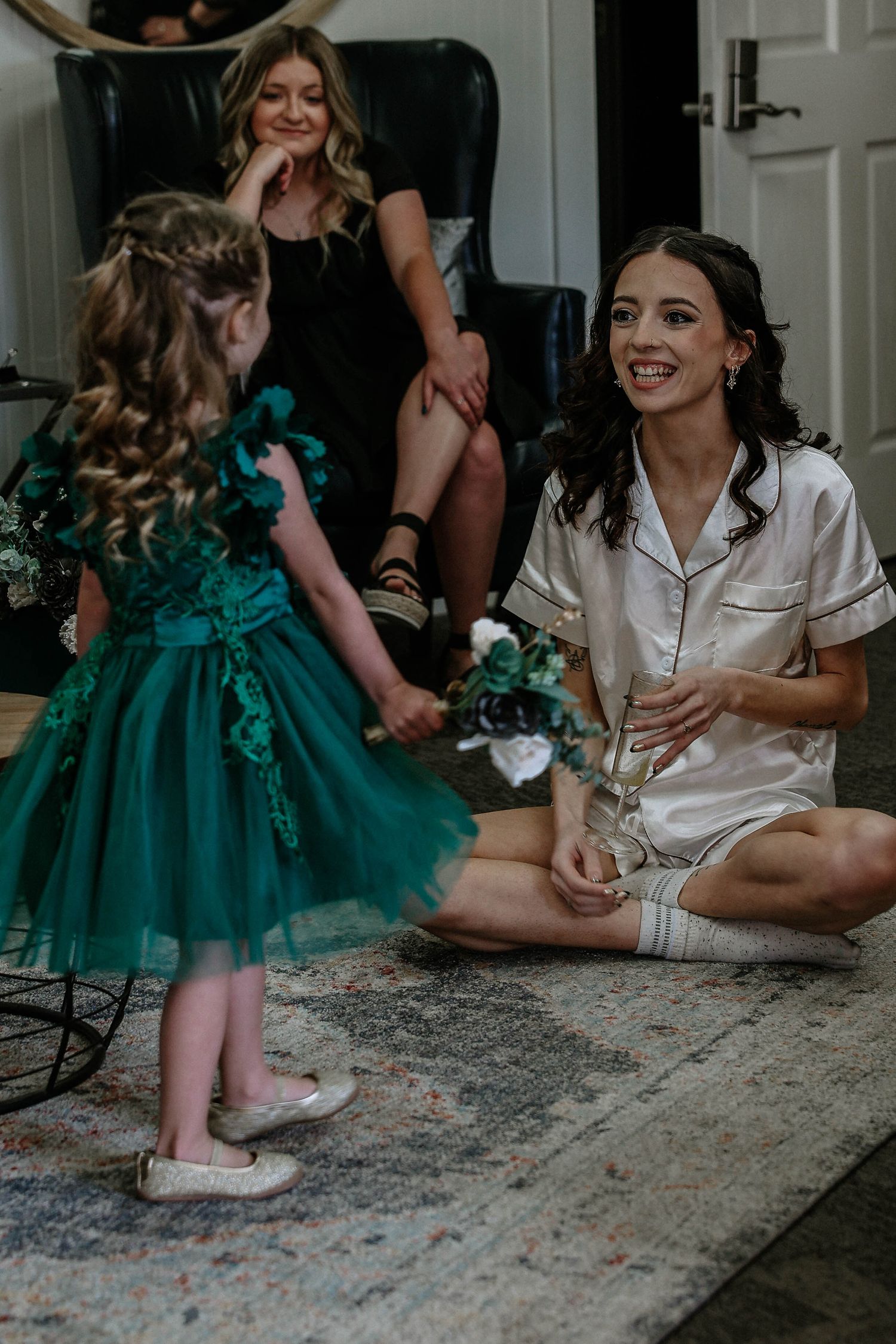 Flower girl in green dress presenting bouquet to smiling bride in white satin pajamas on floor.
