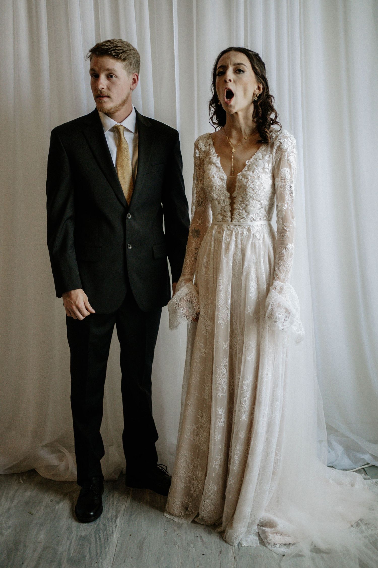 Bride in lace wedding gown and groom in black suit with gold tie posing together against white curtain backdrop.