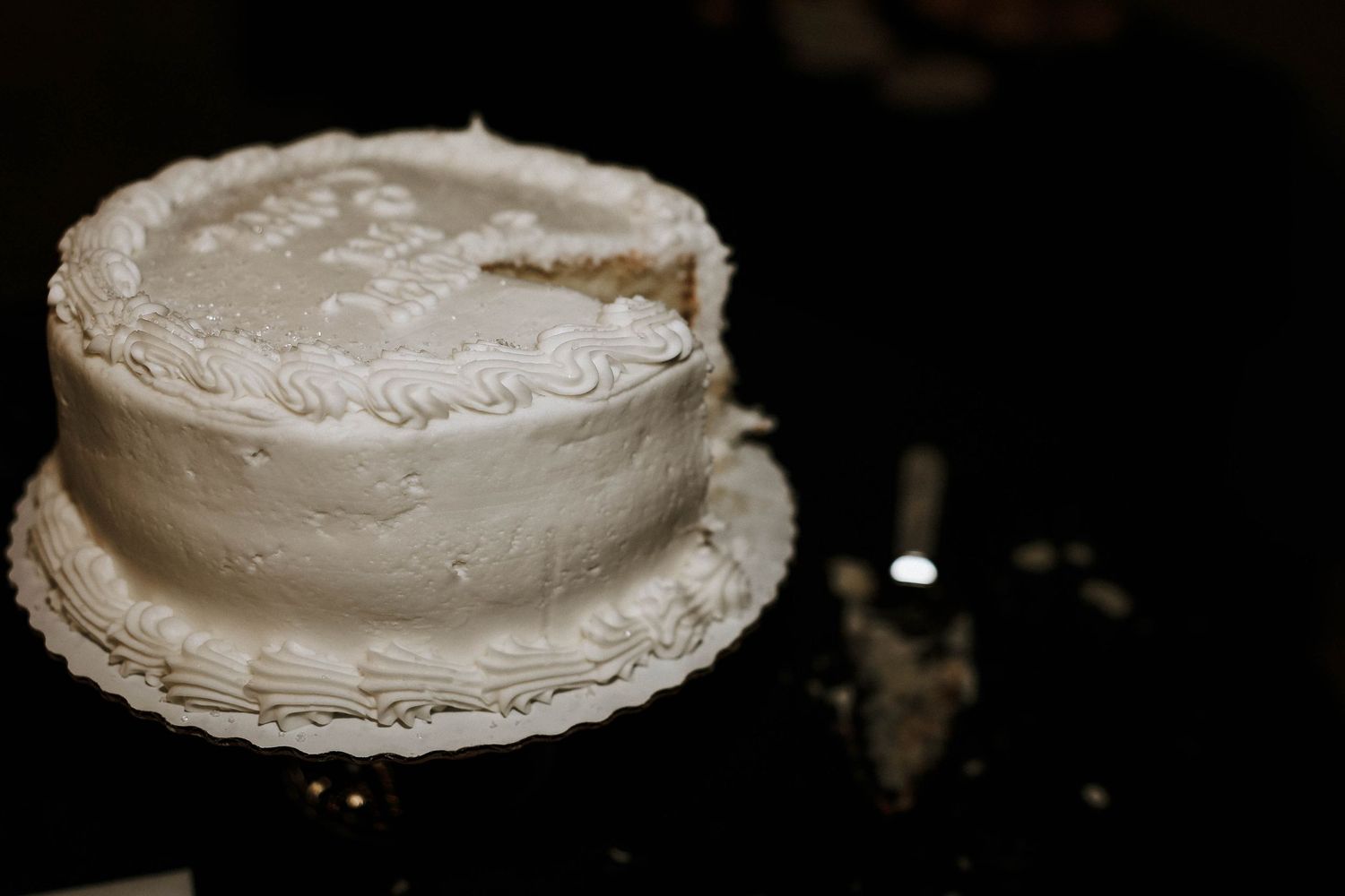 White frosted single-tier wedding cake with decorative piping on a dark reflective surface, partially sliced.