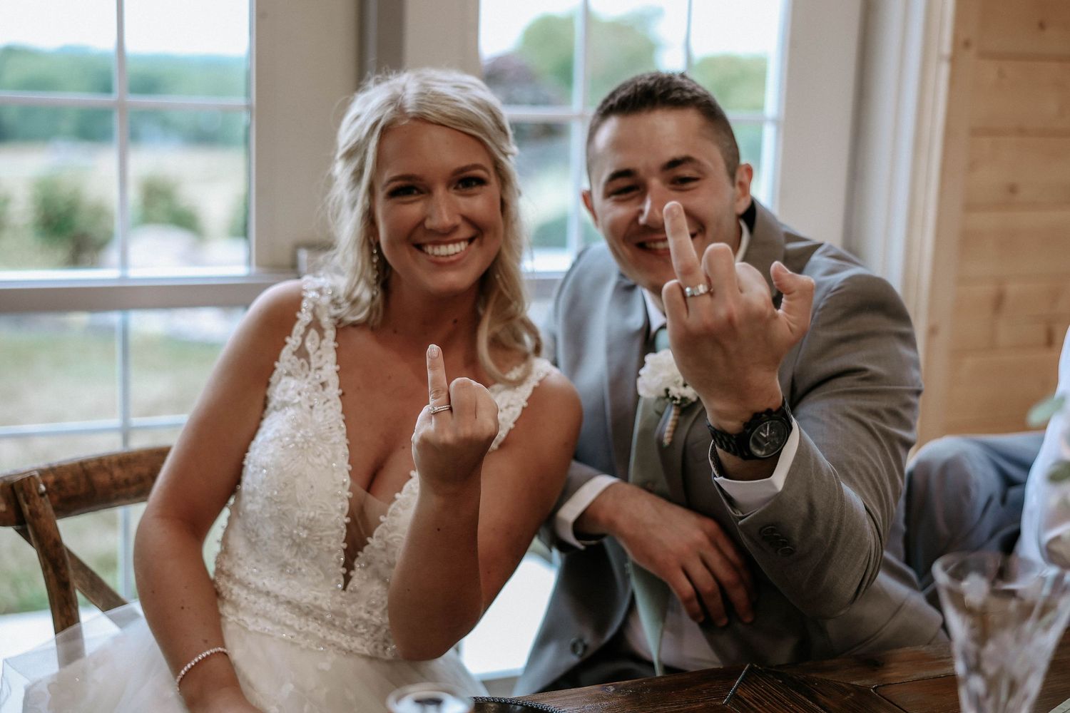 Bride and groom smiling and showing off wedding rings at reception table in rustic venue.