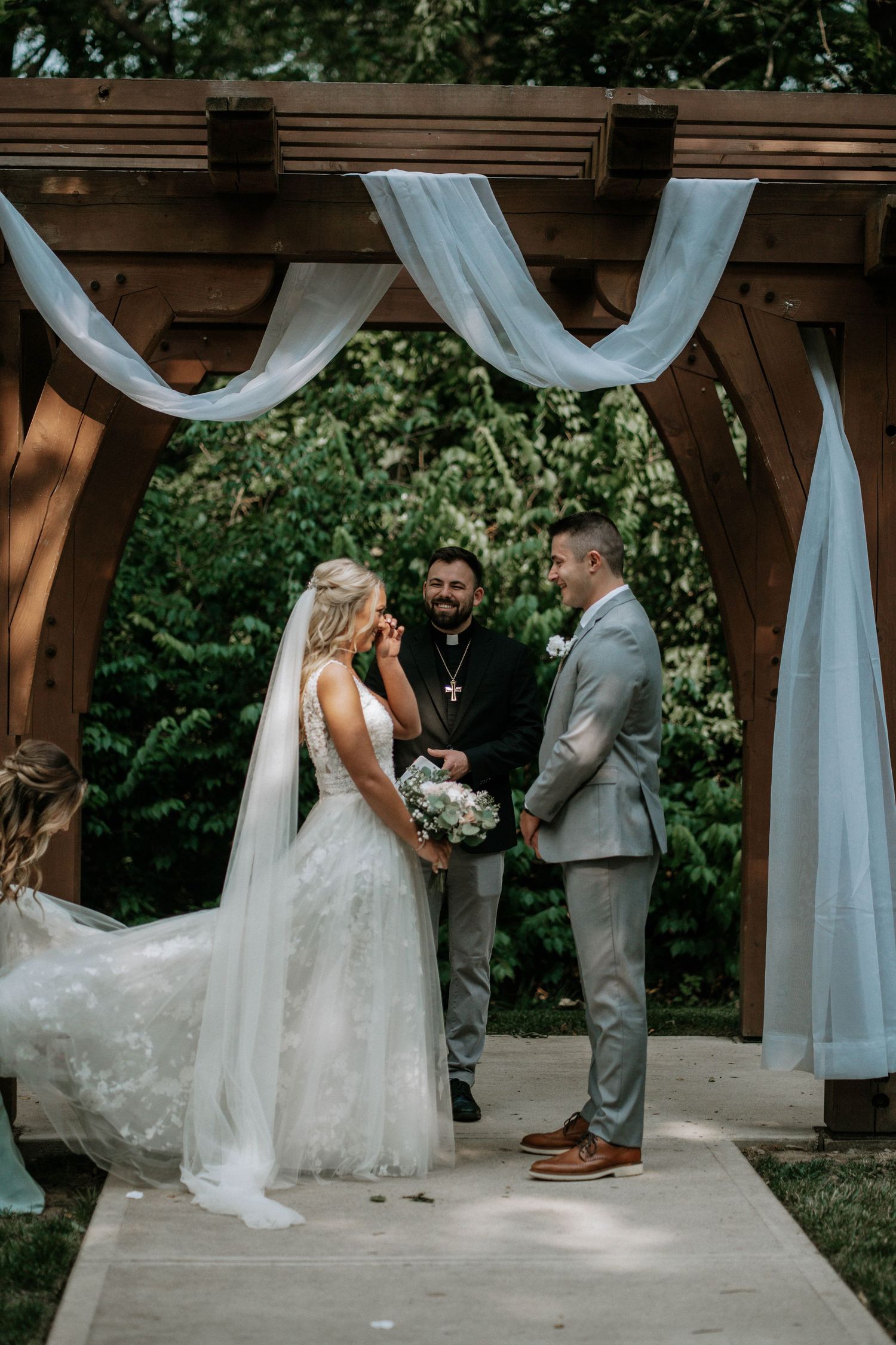 Bride and groom exchange vows under a wooden arch with white draping at an outdoor wedding ceremony.