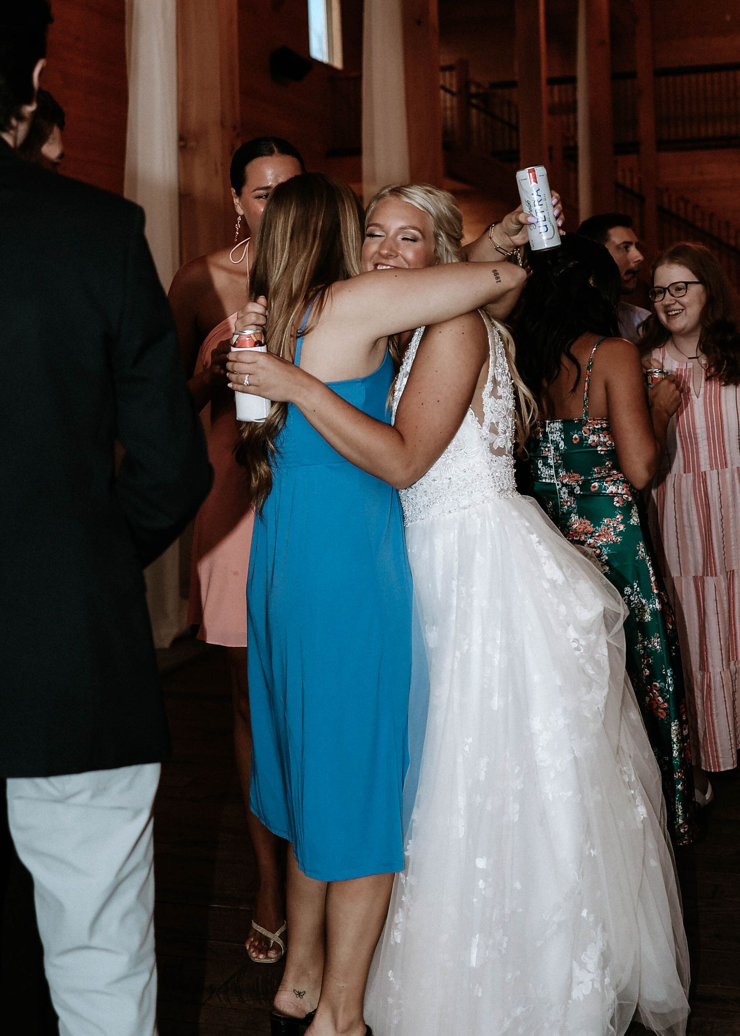 Bride in white lace gown hugging guest in blue dress at wedding reception.