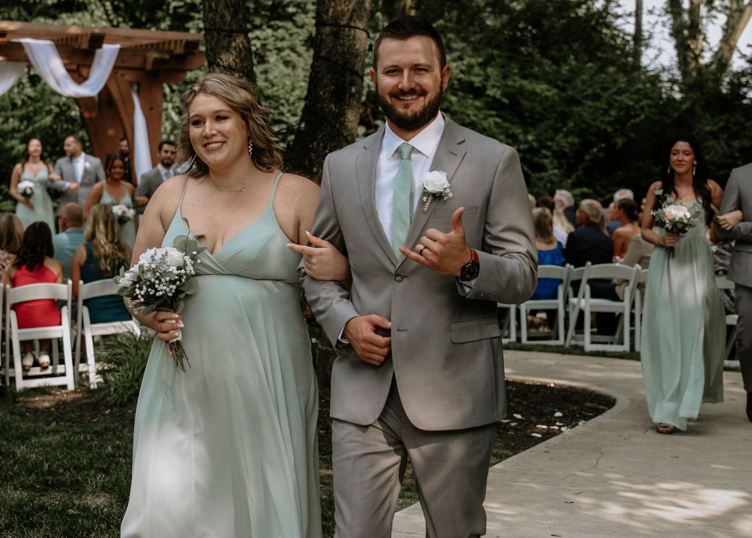 Bridesmaid in mint green gown walks arm-in-arm with groomsman in gray suit at outdoor wedding ceremony.