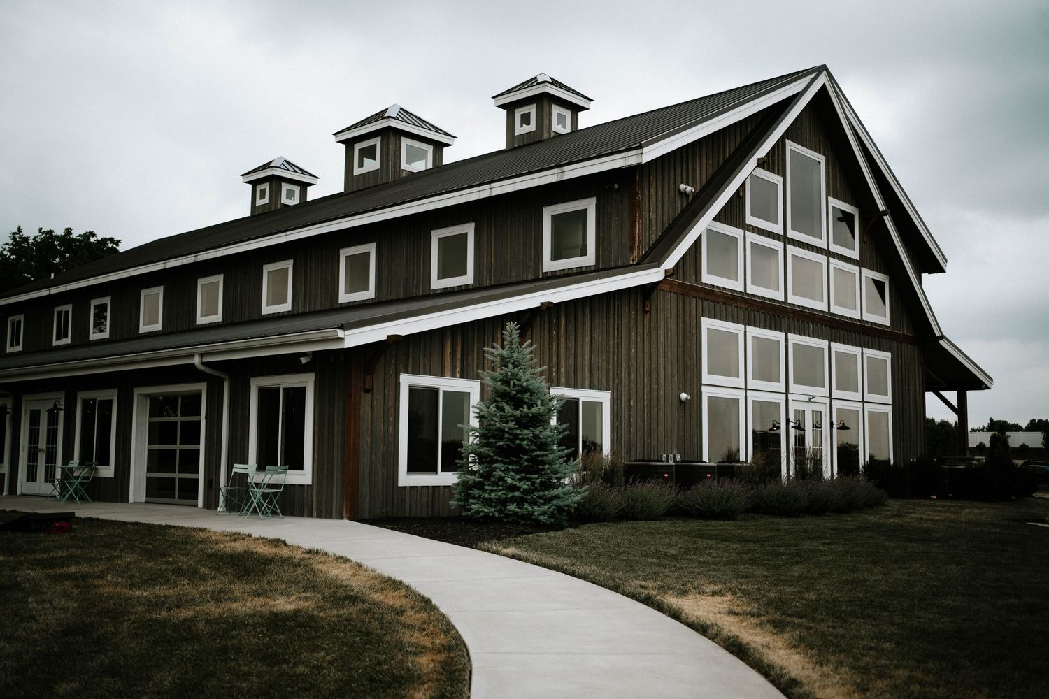 A rustic dark brown barn-style wedding venue with white trim windows and a curved walkway on an overcast day.