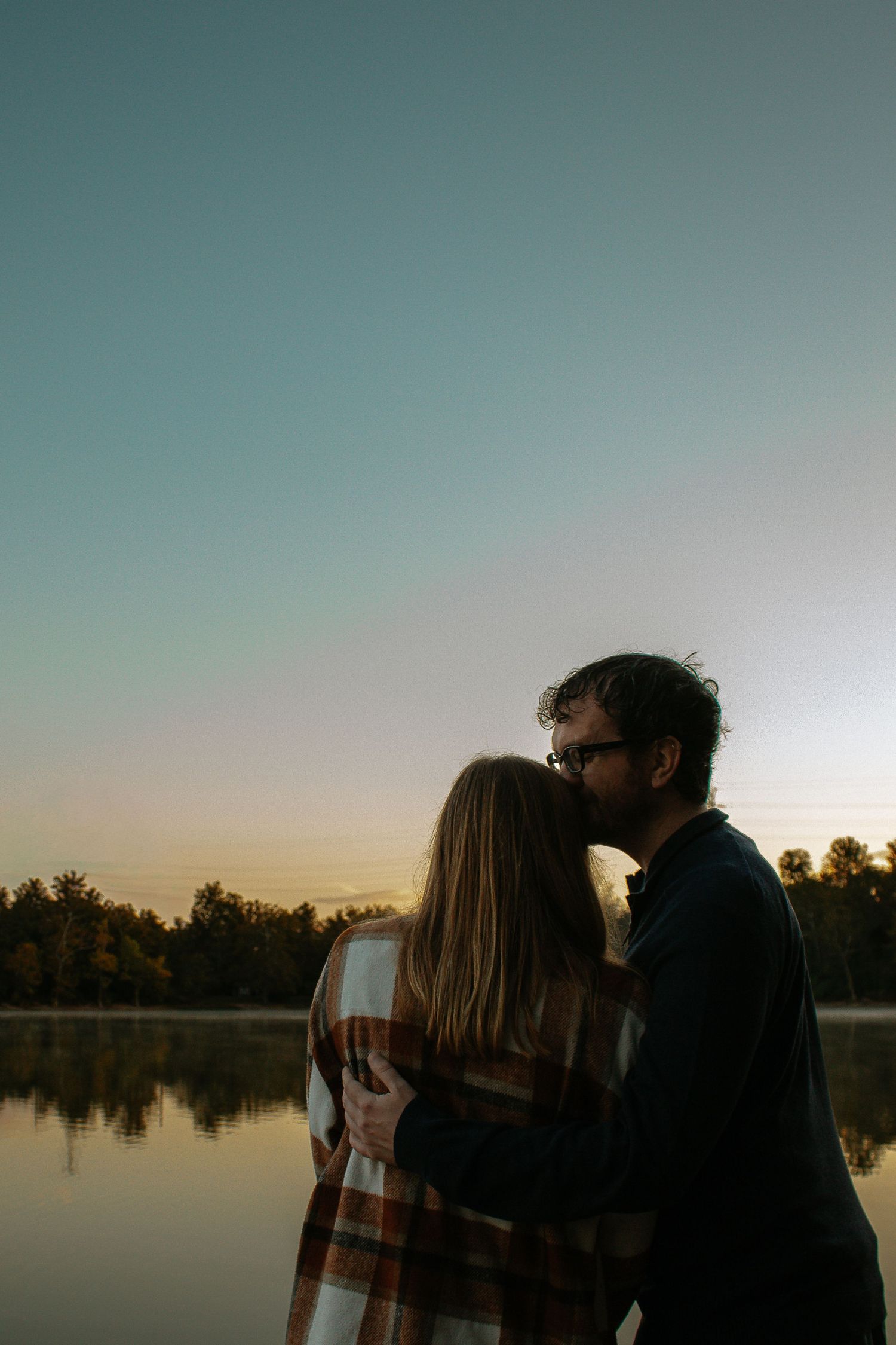 A couple shares a tender kiss by a serene lake at sunset, surrounded by autumn trees in Cincinnati, Ohio