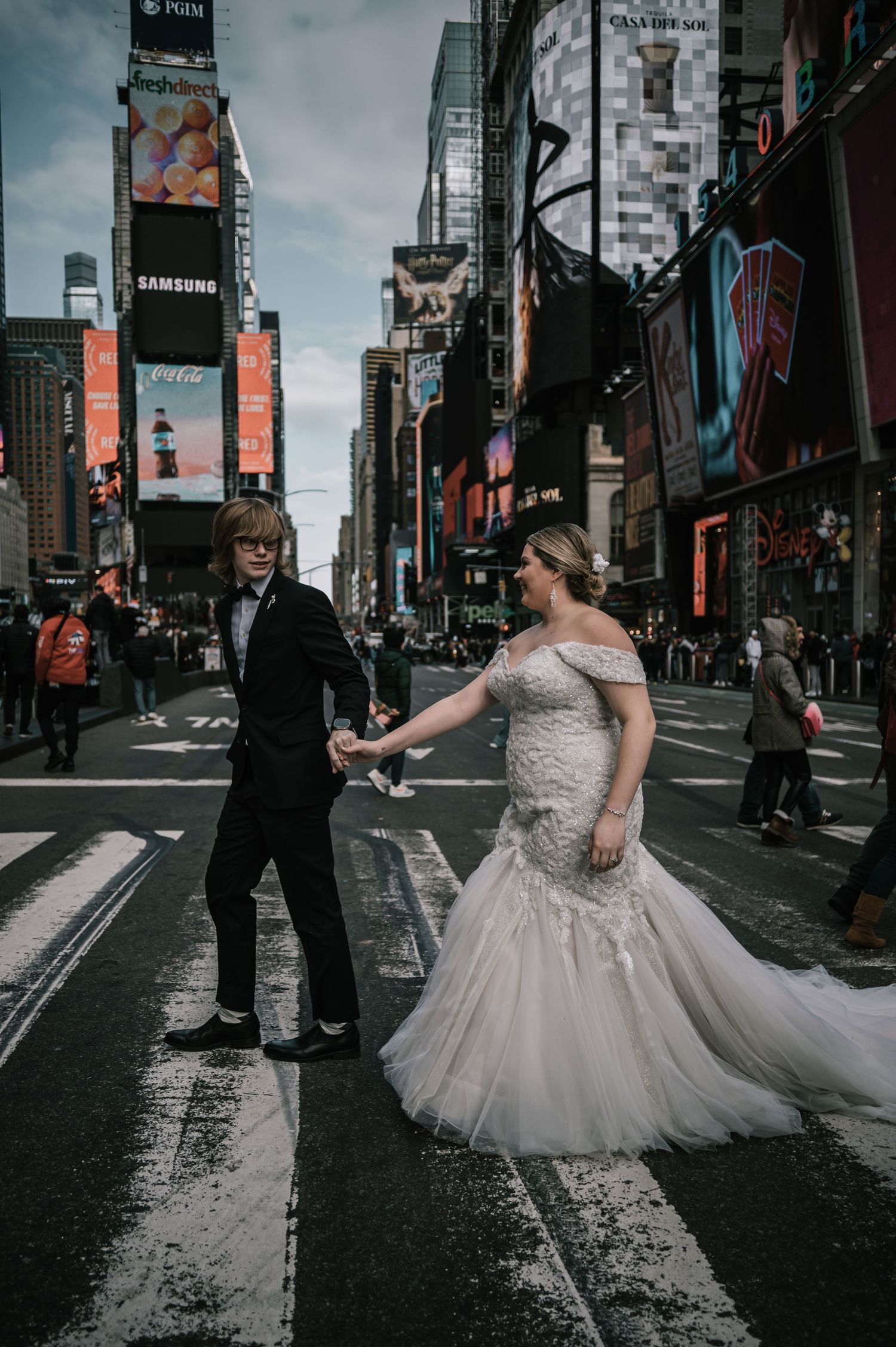 Bride in mermaid gown and groom in tuxedo hold hands crossing Times Square in New York City.
