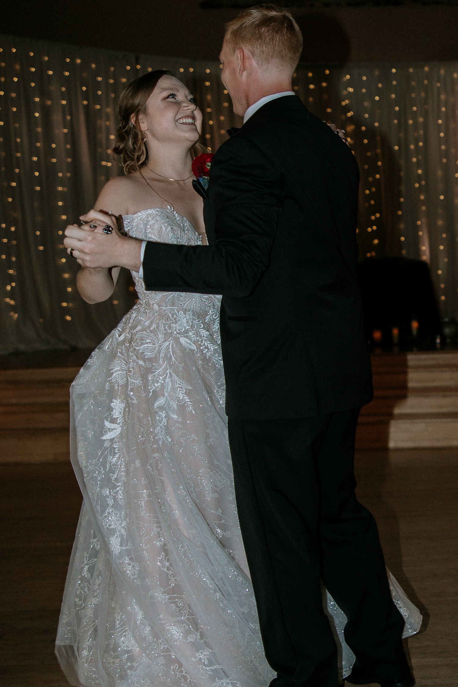 Smiling bride in off-shoulder lace gown dances with groom in black tuxedo at reception at Urban Artifact in Cincinnati, Ohio