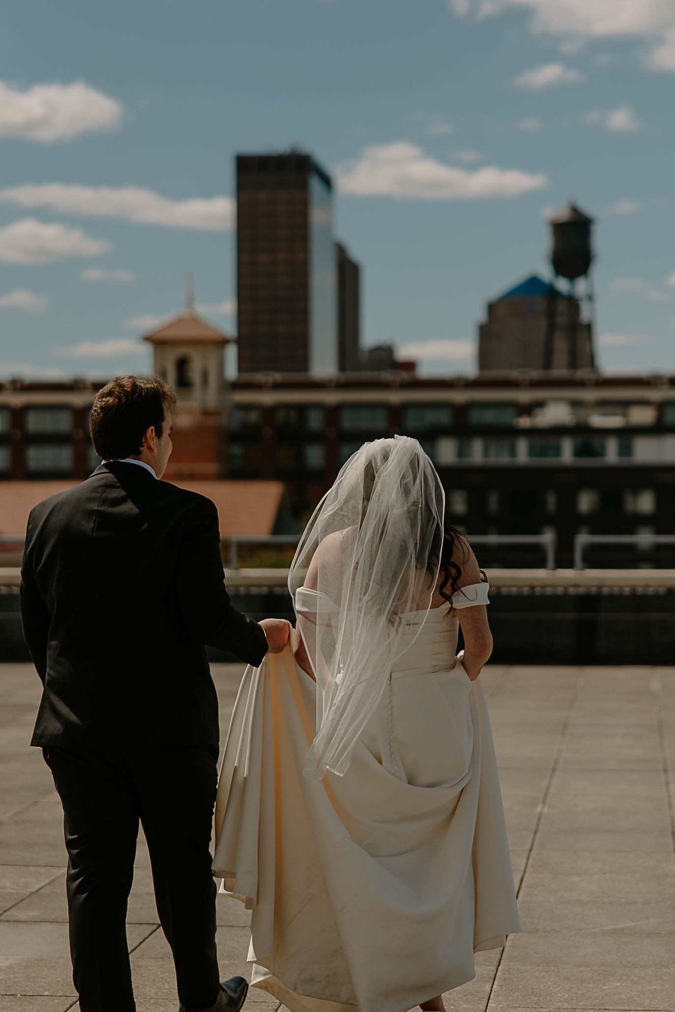 Bride and groom walk hand-in-hand on a rooftop with a city skyline backdrop on their wedding day.