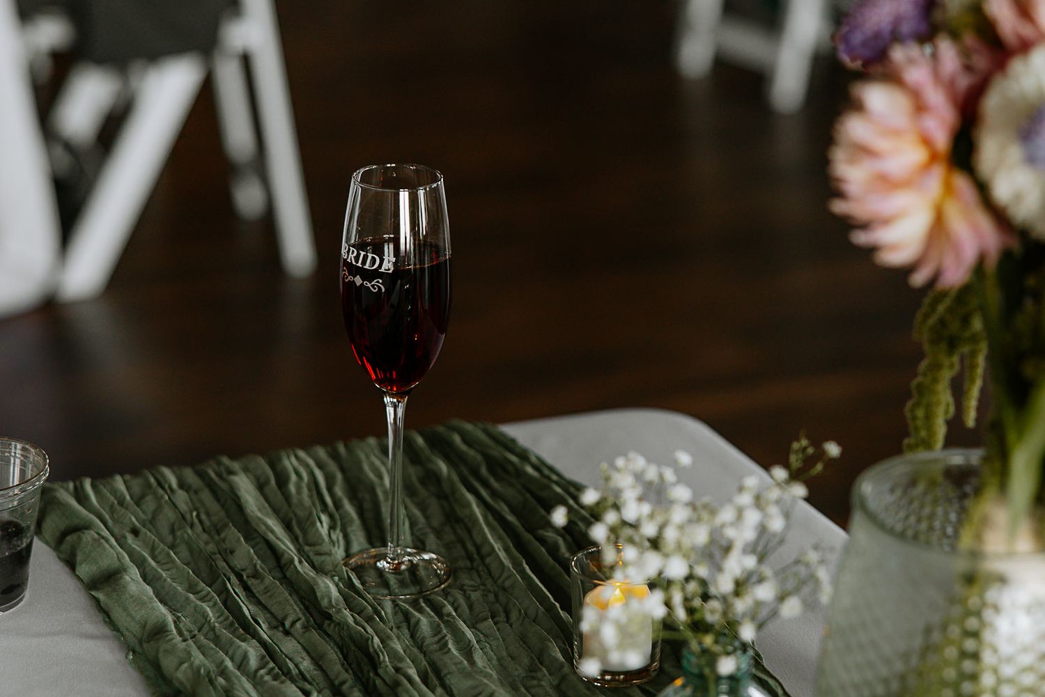 Bride-labeled champagne flute filled with red wine on a green satin wedding table runner.